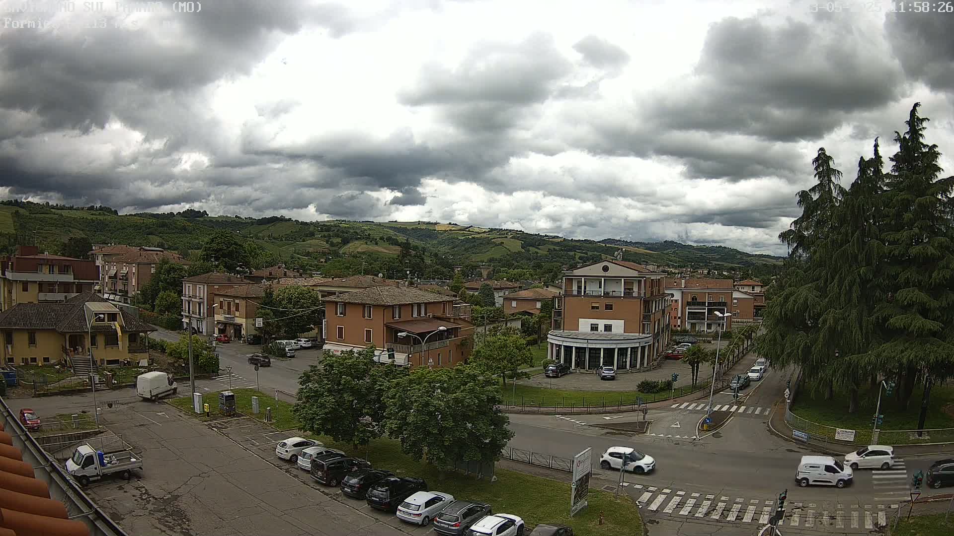 A high-angle view of a town nestled in rolling green hills under a cloudy sky, showing buildings, parked cars, and streets.