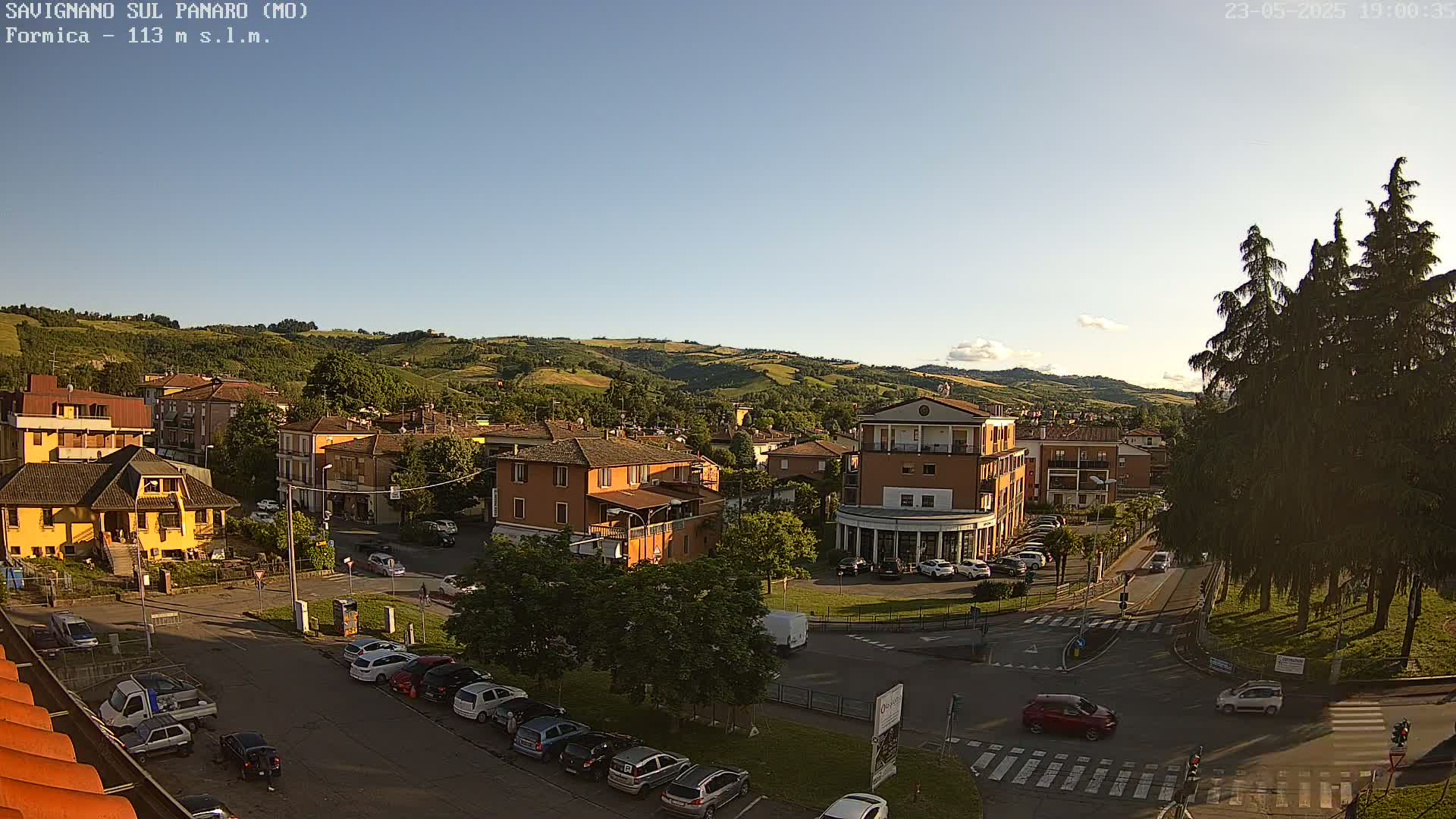 A small Italian town nestled in a valley under a clear, late afternoon sky, with cars parked along the streets and several buildings visible.