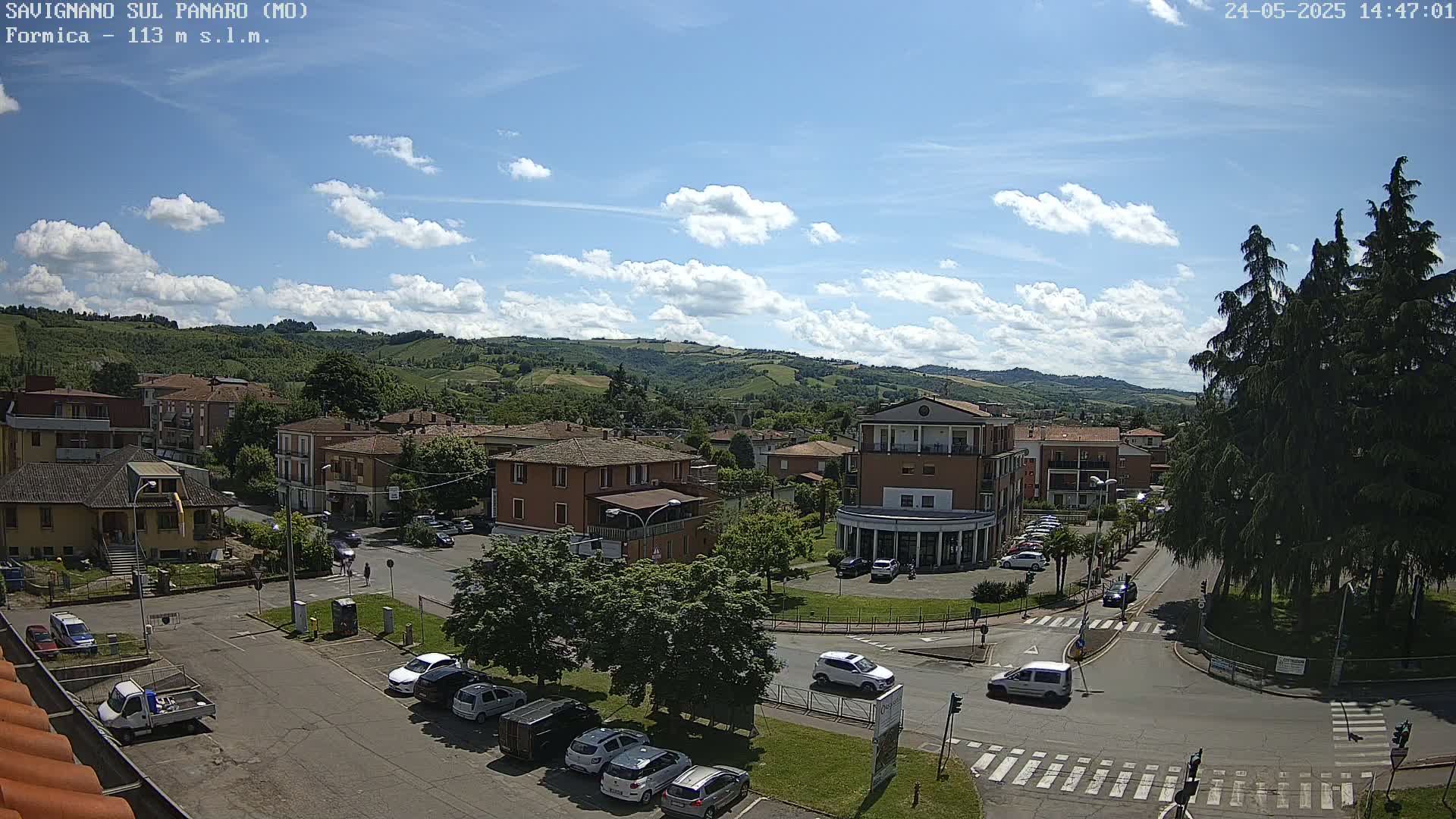 A partly sunny day shows a view of a town nestled in a valley surrounded by rolling green hills, with cars parked in a lot and driving on the roads below.