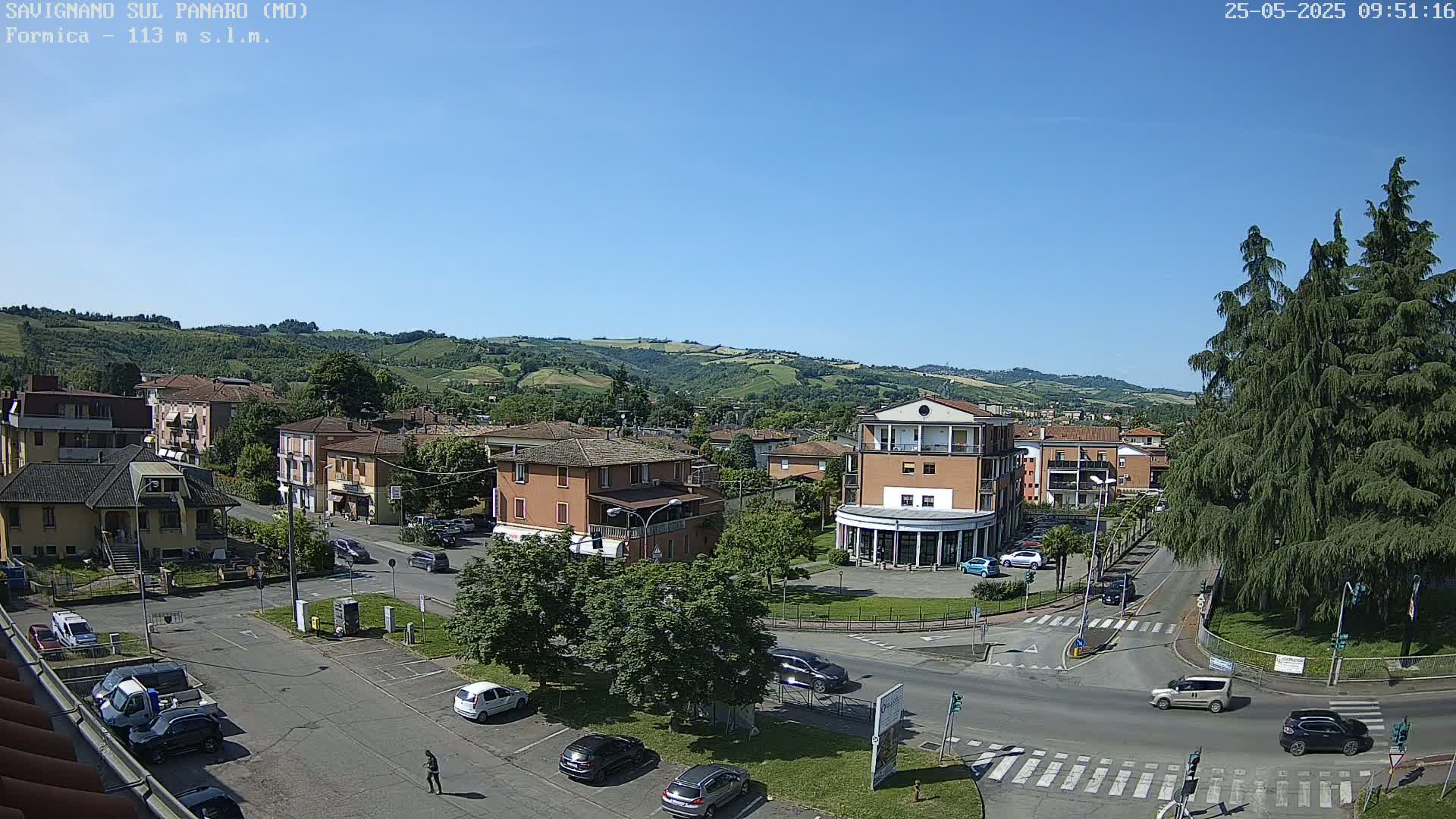 An elevated view of a town square in a small Italian town on a sunny day, surrounded by low buildings and rolling green hills in the background.