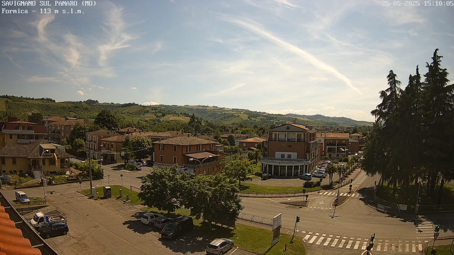 A partly sunny day reveals a small town nestled in a valley, surrounded by rolling green hills, with several cars parked in a lot and along the road.
