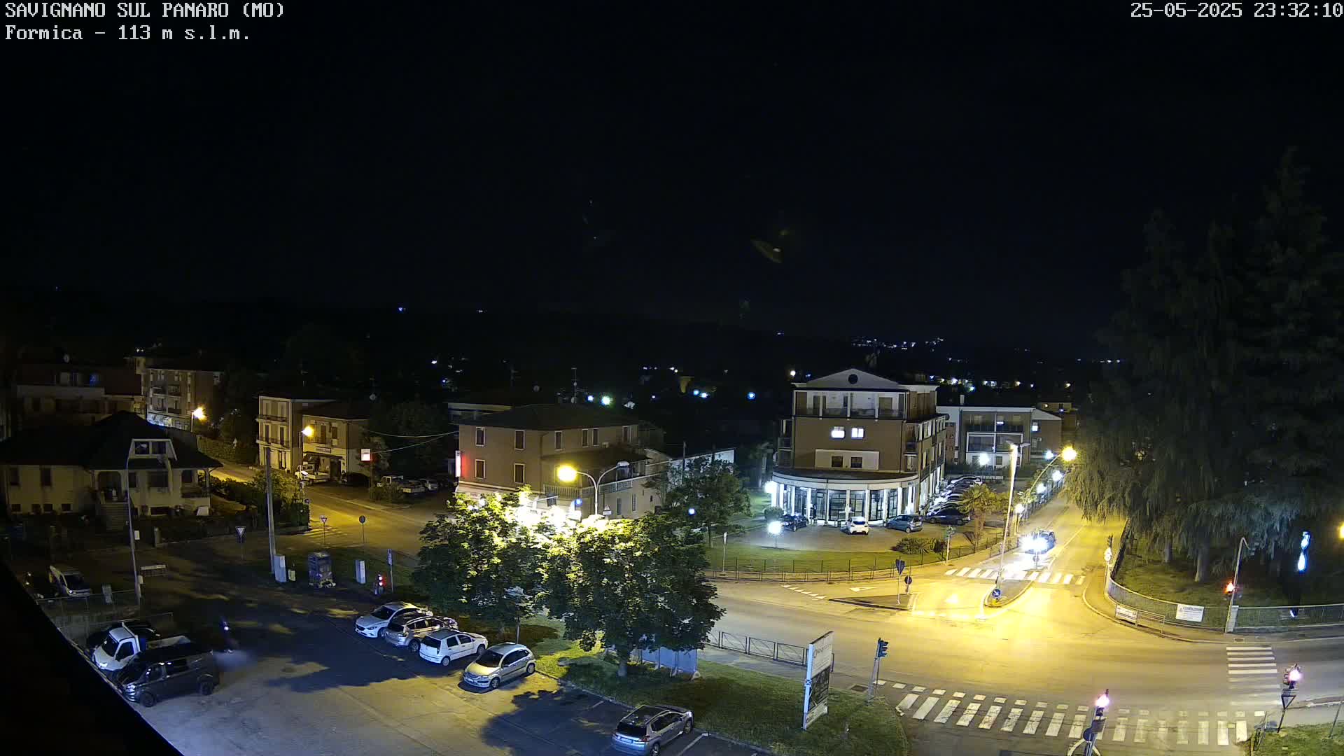 A nighttime aerial view of a town square with several buildings and parked cars under a clear, dark sky.
