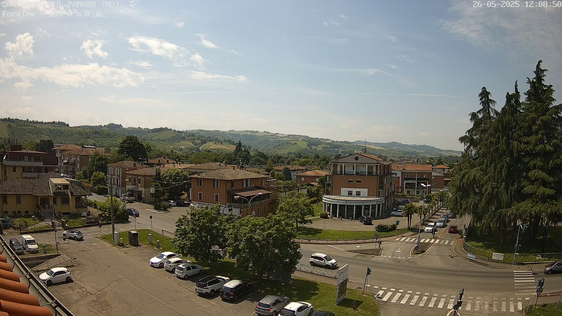 A sunny, partly cloudy day overlooks a town square with several cars parked and driving, surrounded by low-rise buildings and green hills in the distance.