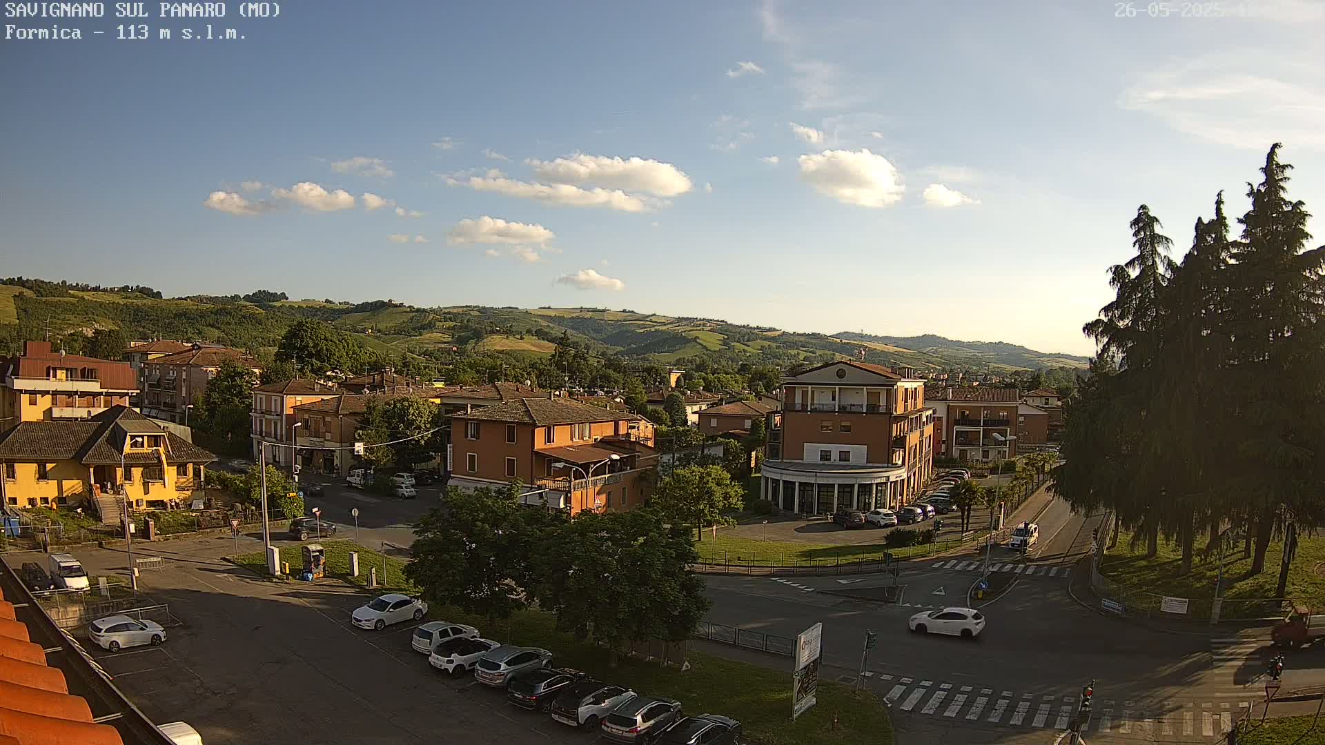 A sunny day with some clouds over a town nestled in rolling green hills, showing a high-angle view of buildings, streets, cars, and trees.