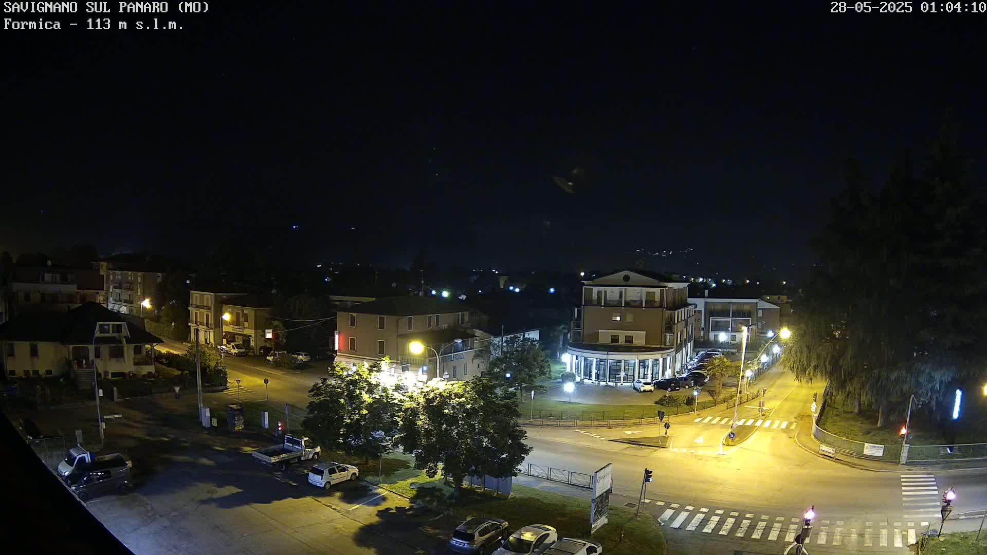 A nighttime overhead view of a town square with several buildings and cars, illuminated by streetlights under a clear, dark sky.