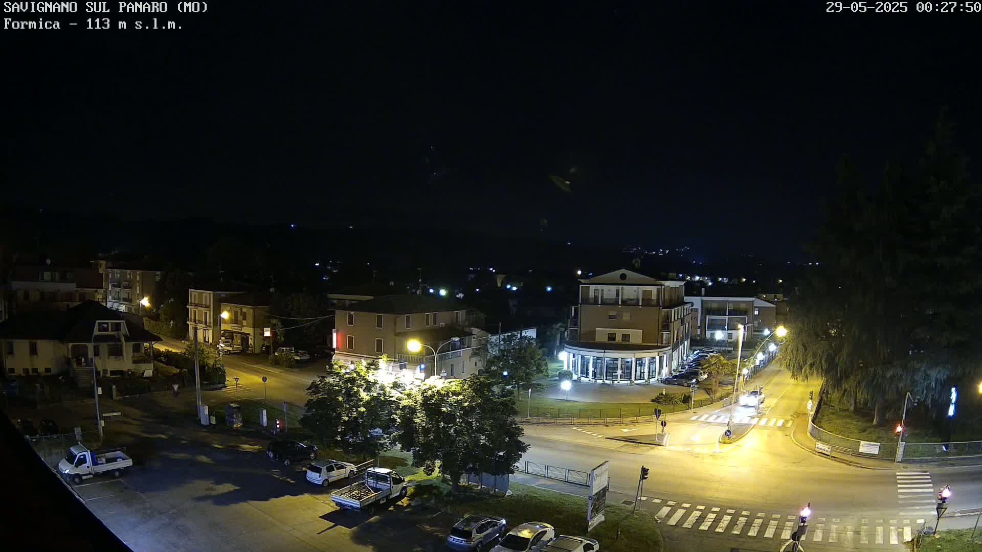 A nighttime, high-angle view of a town square with several buildings, cars, and streetlights illuminated under a clear, dark sky.