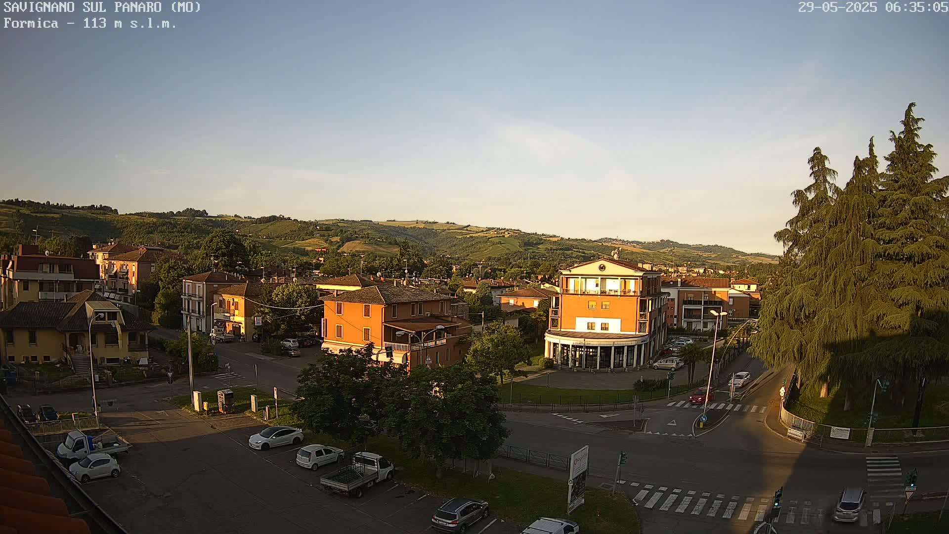 A high-angle view of a town nestled in rolling hills under a clear, sunset sky, showing a street intersection with cars and buildings.