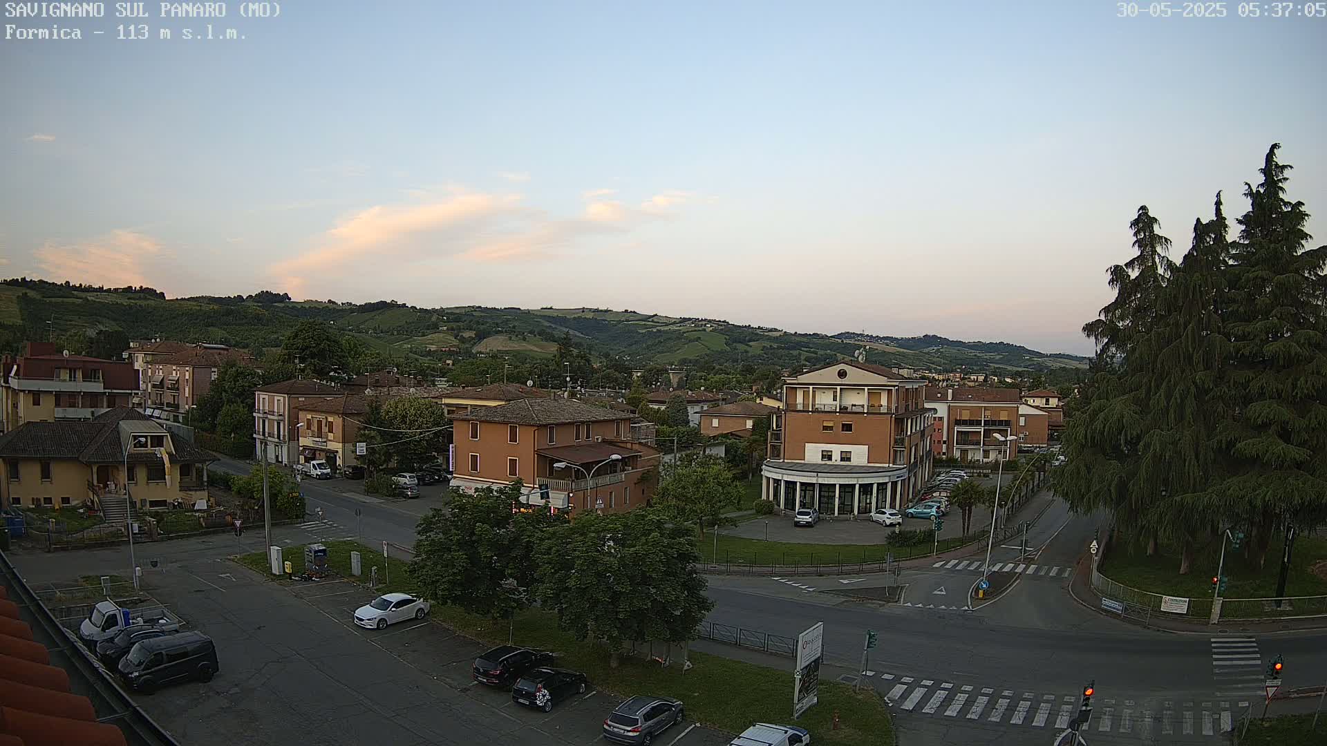 A mostly clear, twilight sky overlooks a town nestled in a valley of rolling green hills, with cars parked in a lot and driving on streets below.