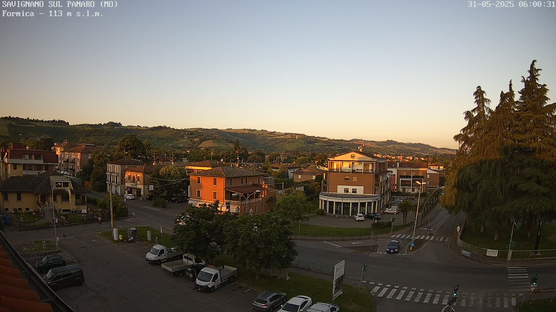 A high-angle, long shot of a town at sunset, with cars parked in a lot and on the road, and a few cars driving, situated before hills under a clear sky.