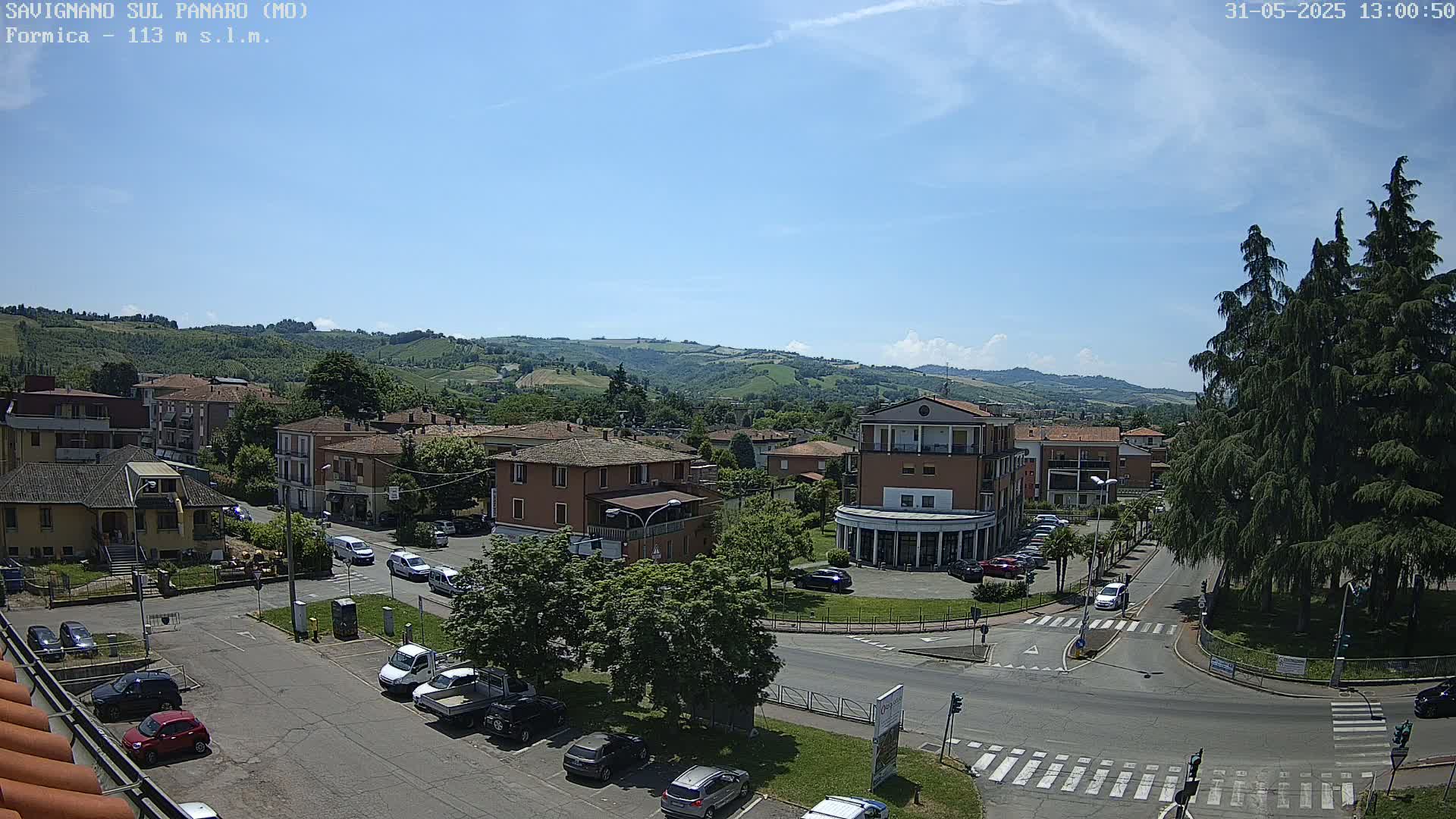 A sunny, high-angle view of a town square with parked cars, buildings, and lush green hills in the background.