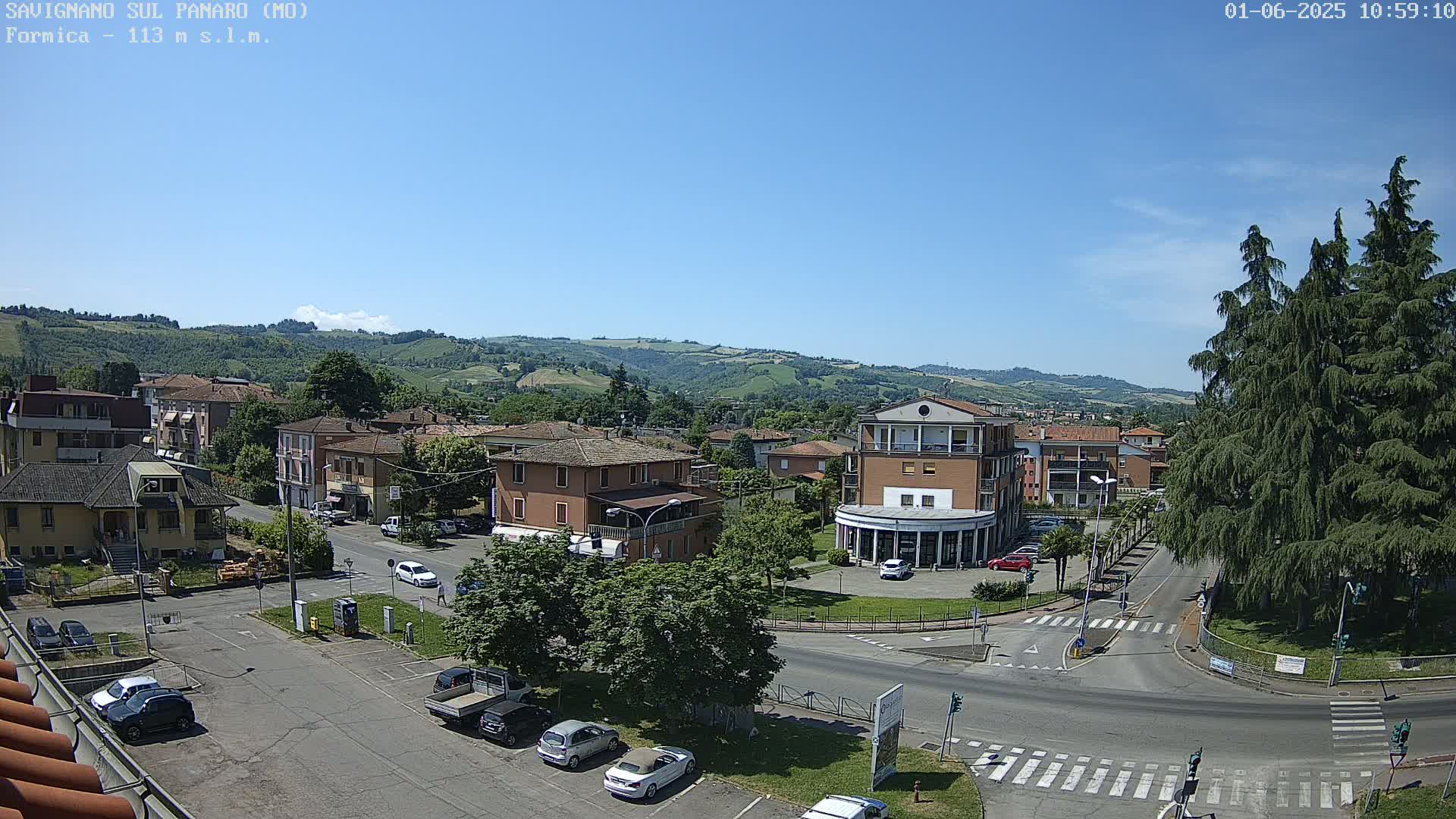 A sunny, high-angle view of a town nestled in a valley, surrounded by green hills and featuring a central intersection with cars and buildings.