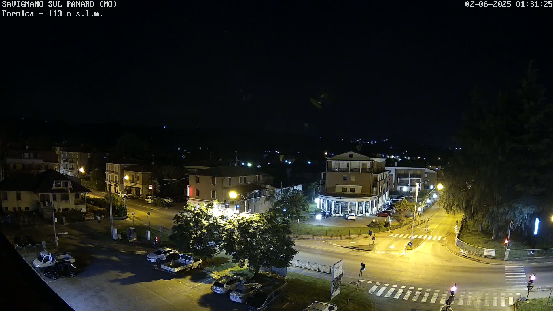 A nighttime aerial view of a town square with several buildings, cars in parking lots, and streetlights illuminating the scene under a clear, dark sky.