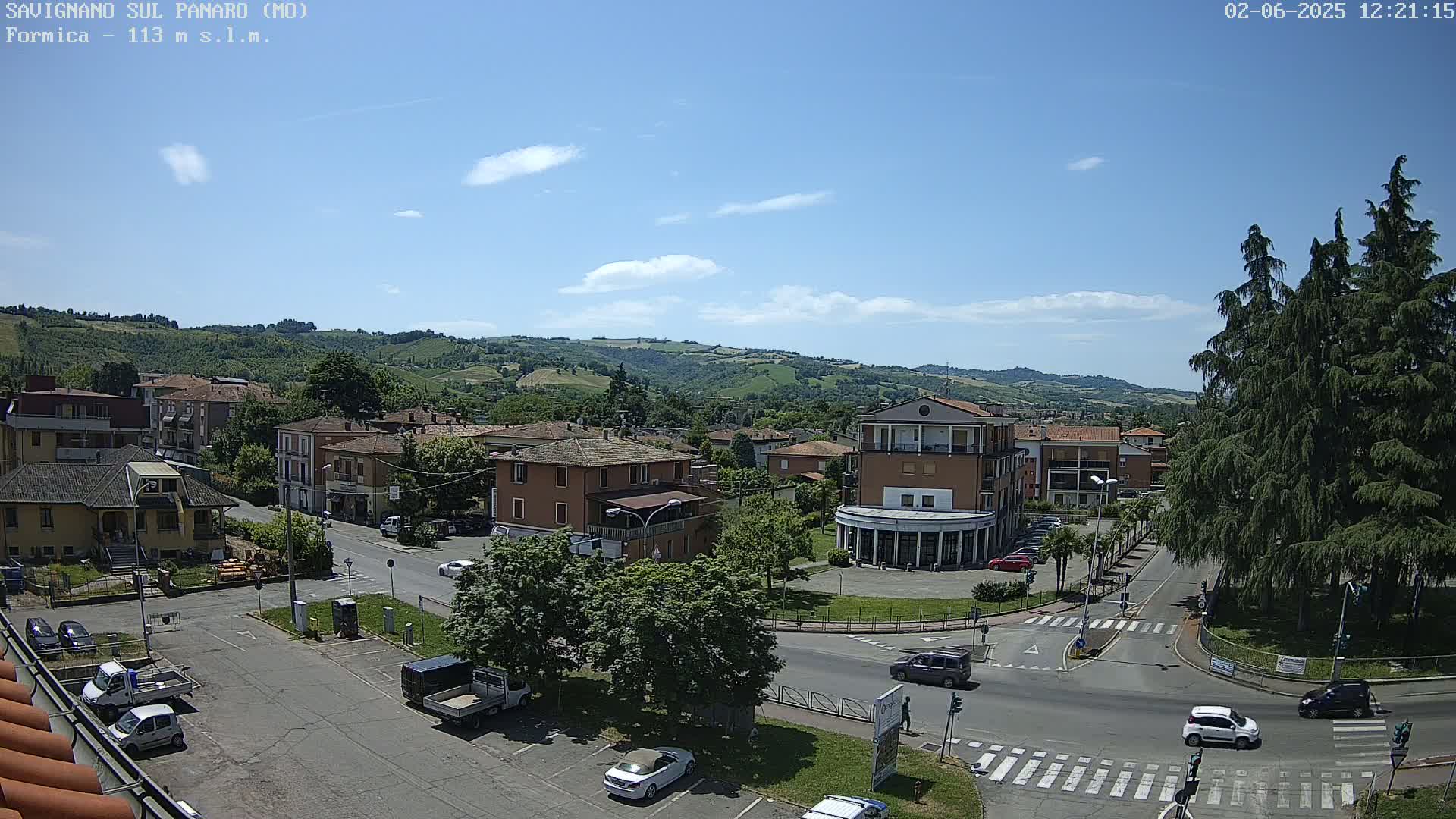 A sunny day reveals a view of a town square, surrounded by low-rise buildings and lush green hills in the background.