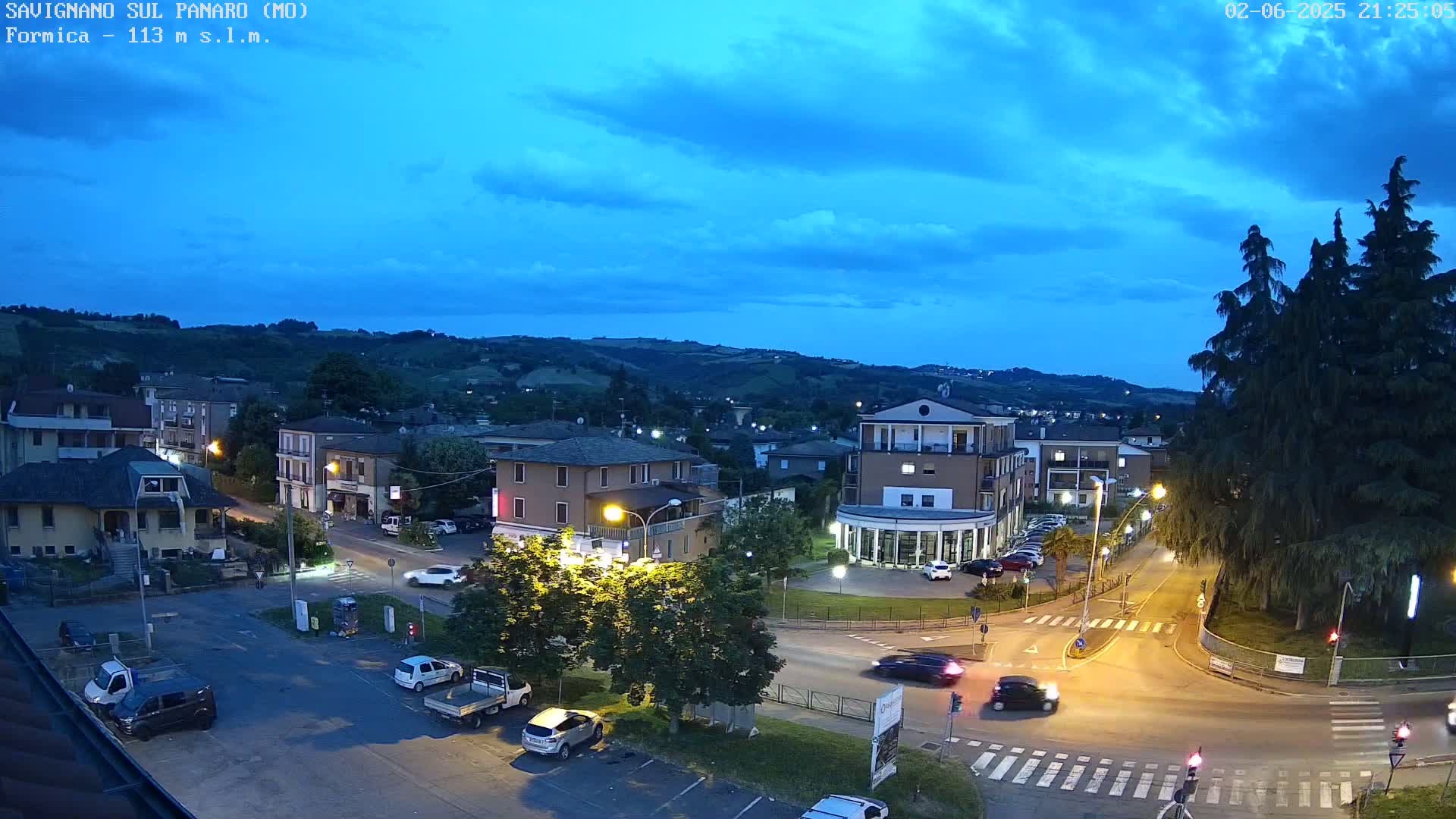A nighttime view of a town nestled in a valley under a partly cloudy, twilight sky, with cars driving on roads that intersect near a central square.