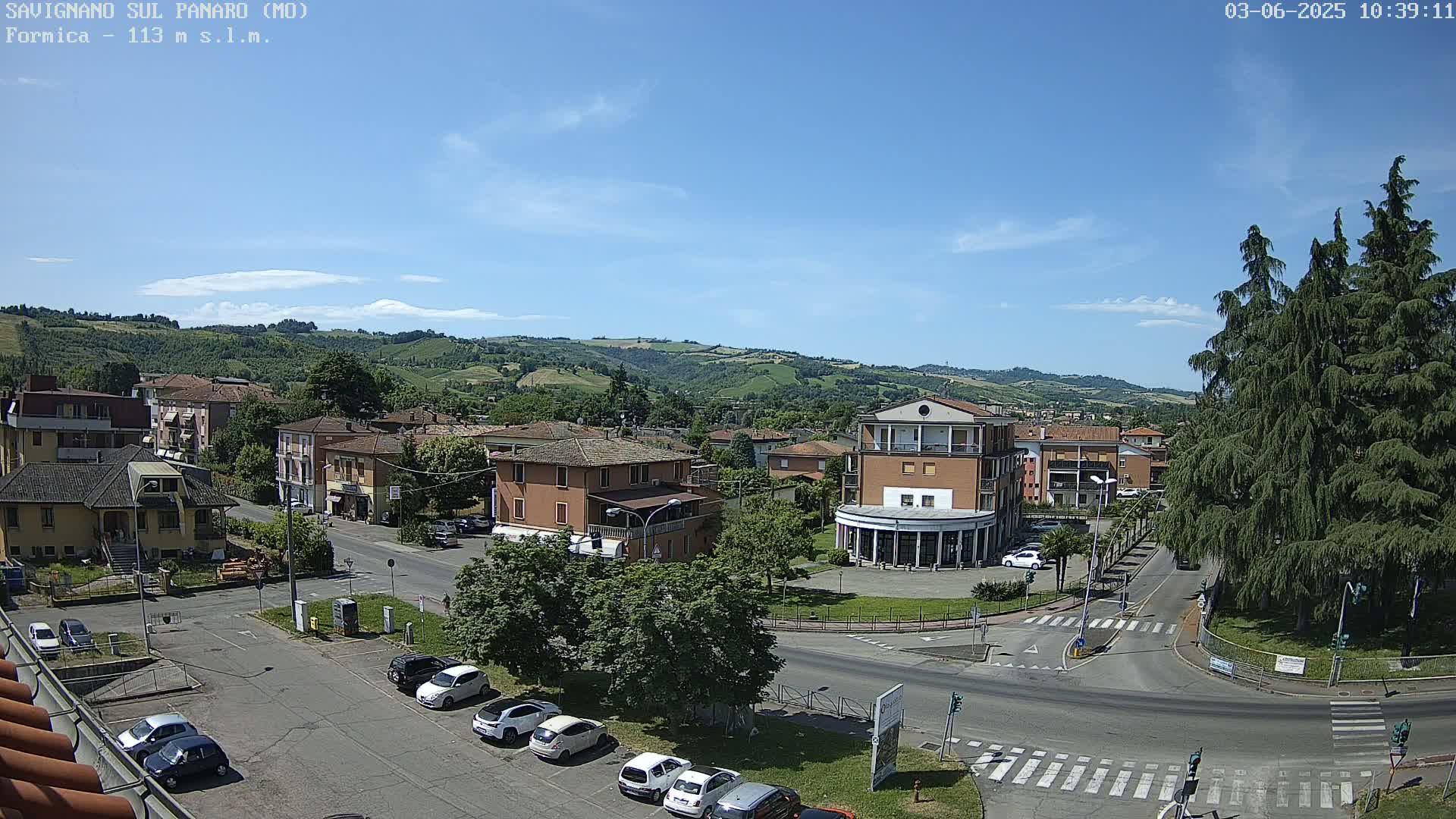 A sunny day reveals a high-angle view of a town nestled in a valley, with cars parked in lots and roads intersecting near a small plaza, surrounded by buildings and greenery.