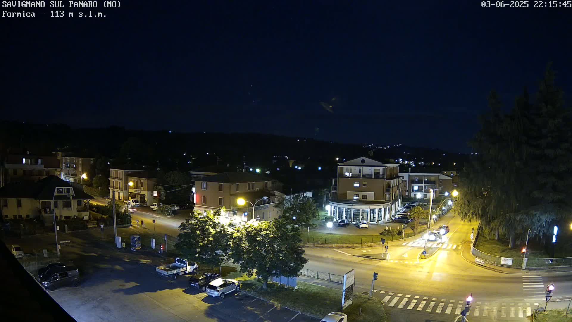 A nighttime, overhead view of a town square with cars parked and driving on roads that intersect, surrounded by buildings and trees under a clear, dark sky.