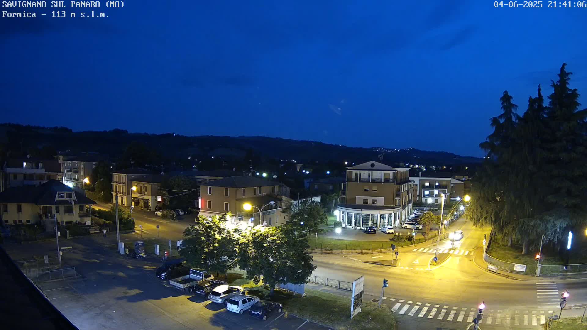 A nighttime, high-angle view of a town square with cars parked and driving on roads illuminated by streetlights, under a dark blue twilight sky.