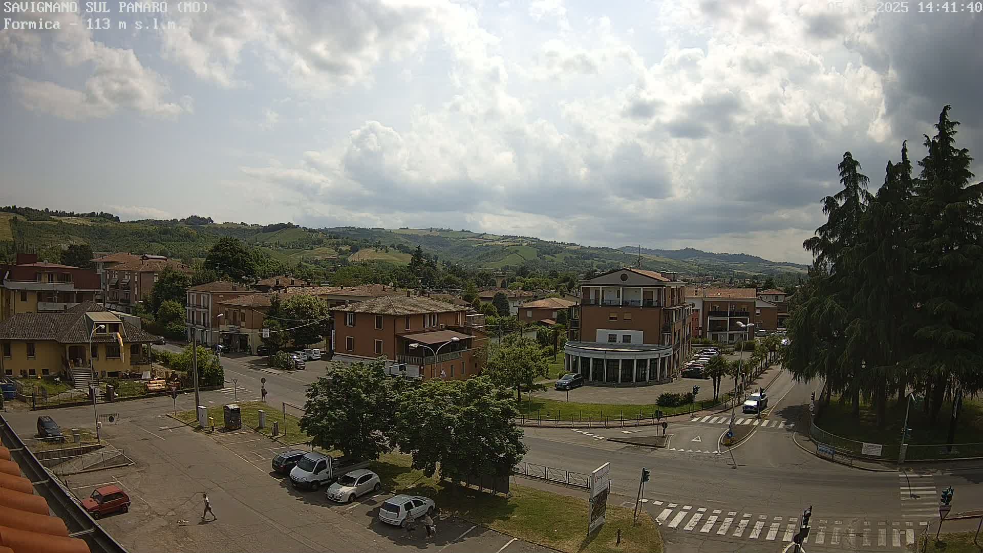 A partly cloudy day reveals a small town nestled in a valley, surrounded by rolling green hills and several cars parked in a lot.