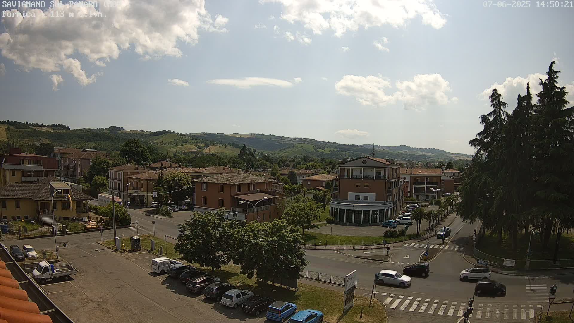 A partly cloudy sunny day overlooks a town nestled in a valley, with cars parked along streets and buildings surrounding a small roundabout.
