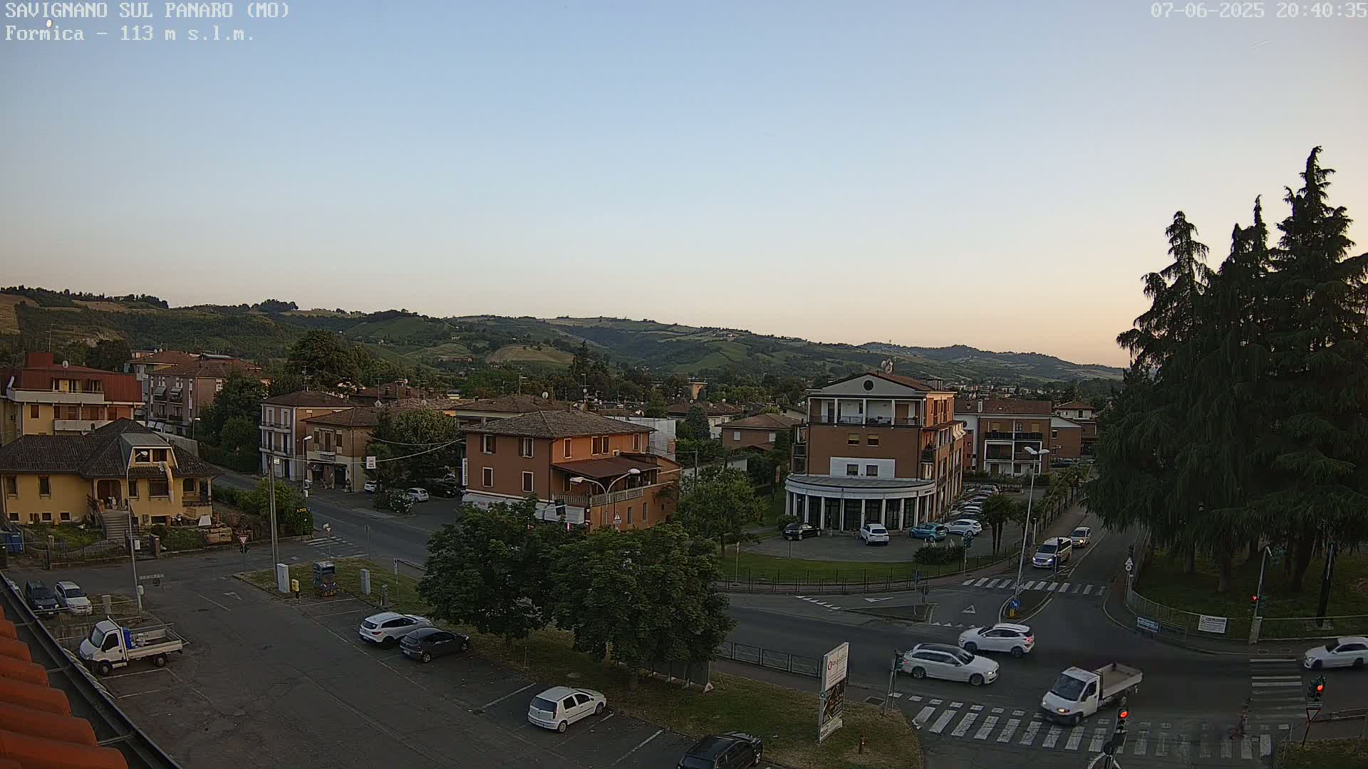A high-angle, long shot of a town square with cars parked and driving around it at dusk, under a clear, light-blue sky.