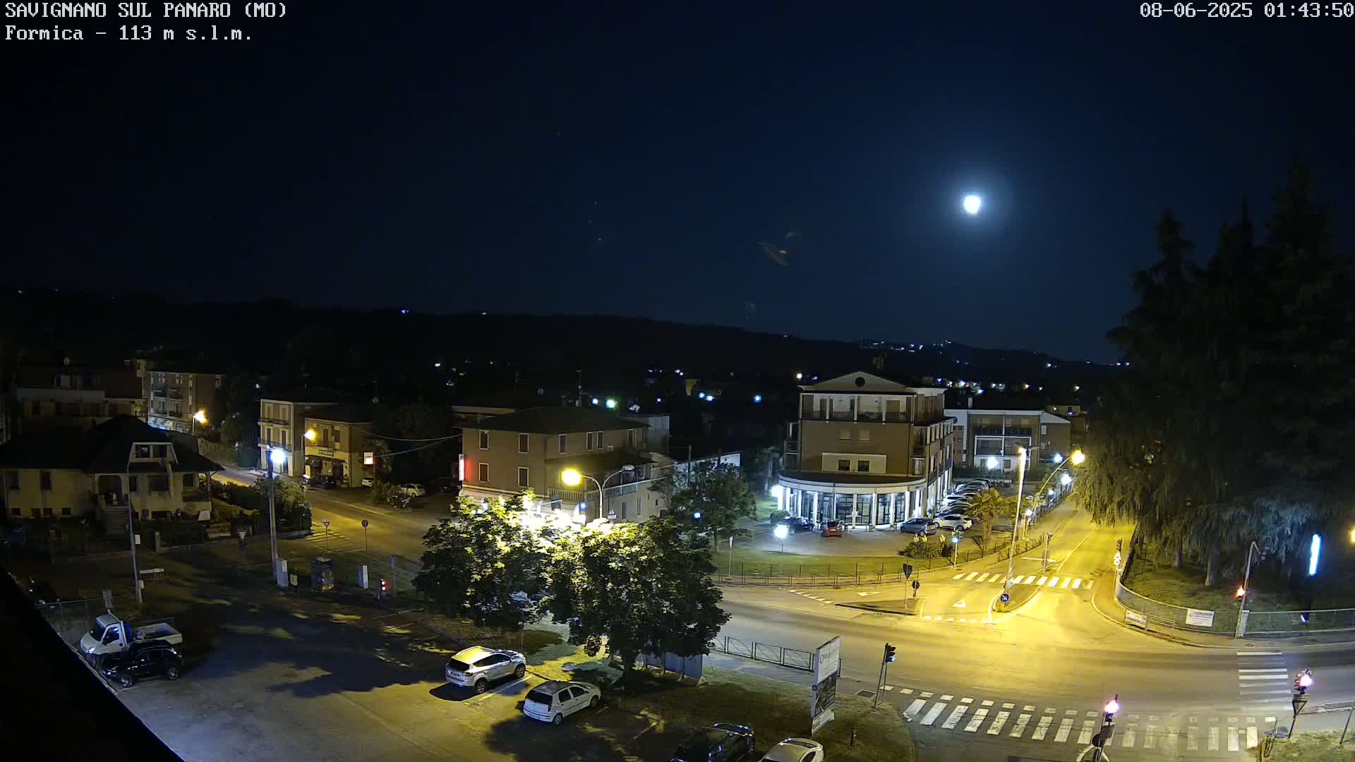 A nighttime, high-angle view of a town square with streetlights illuminating cars parked along the roadways under a bright moon.