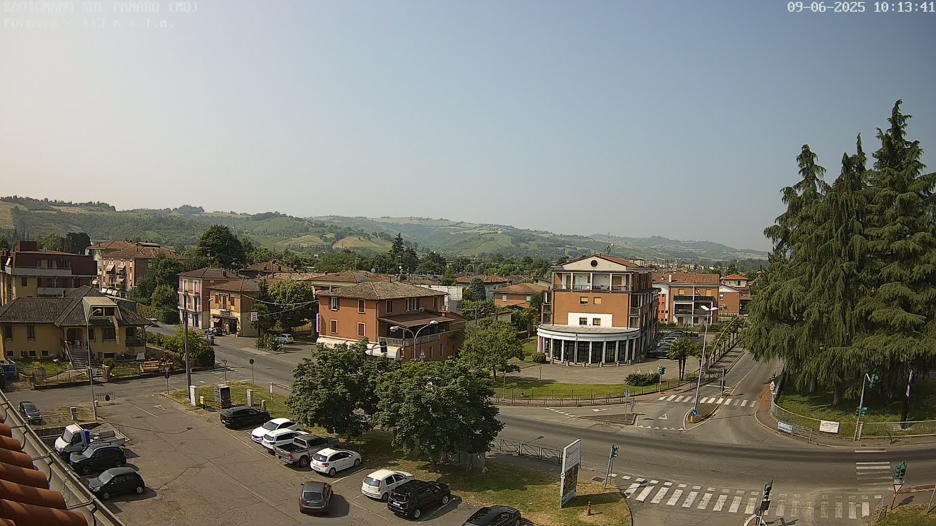 A sunny day reveals a town square with parked cars, surrounded by buildings and rolling green hills in the background.