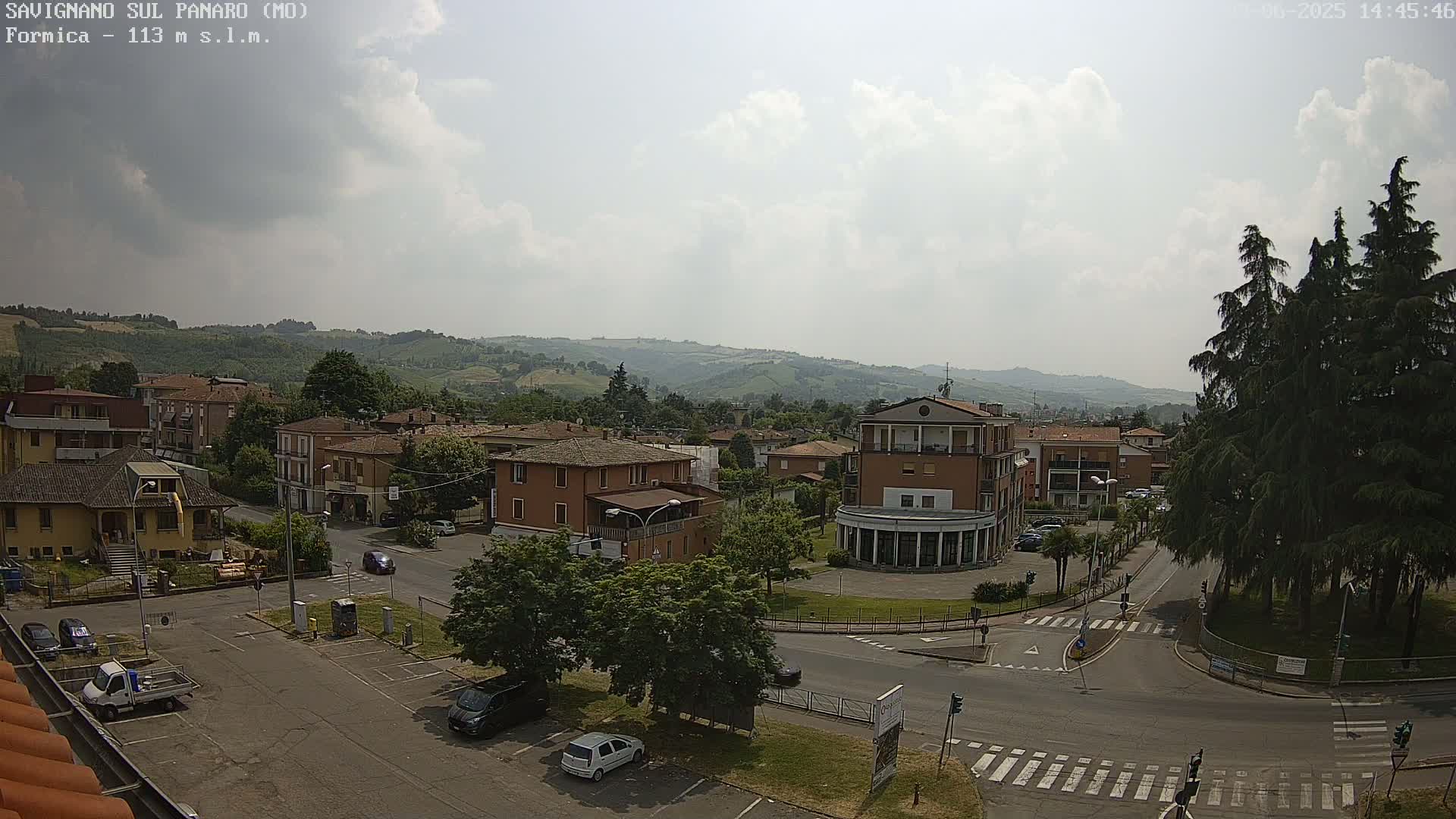 A partly cloudy day reveals a view of a town nestled in a valley, with buildings, cars, and trees visible, and rolling hills in the background.