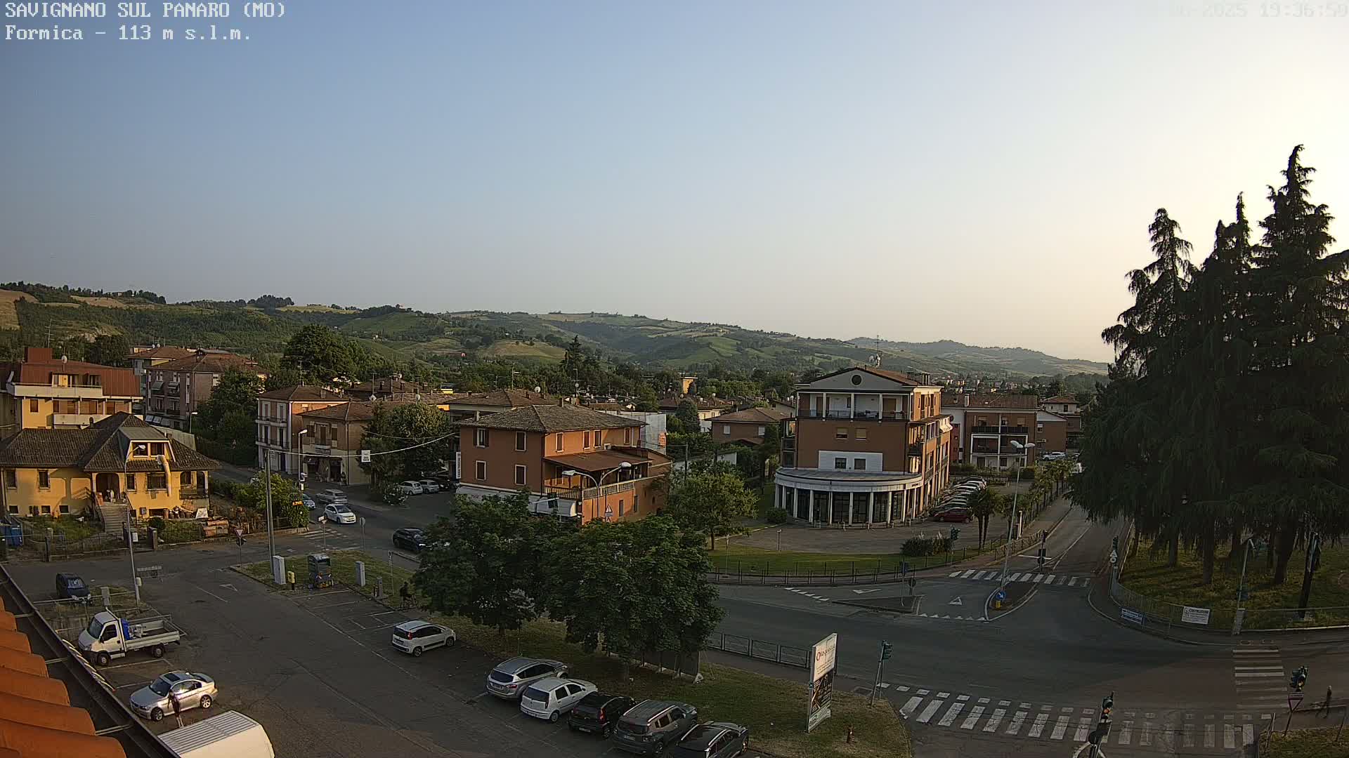 A high-angle, long shot shows a town square with several buildings and parked cars under a clear, light-blue sky.