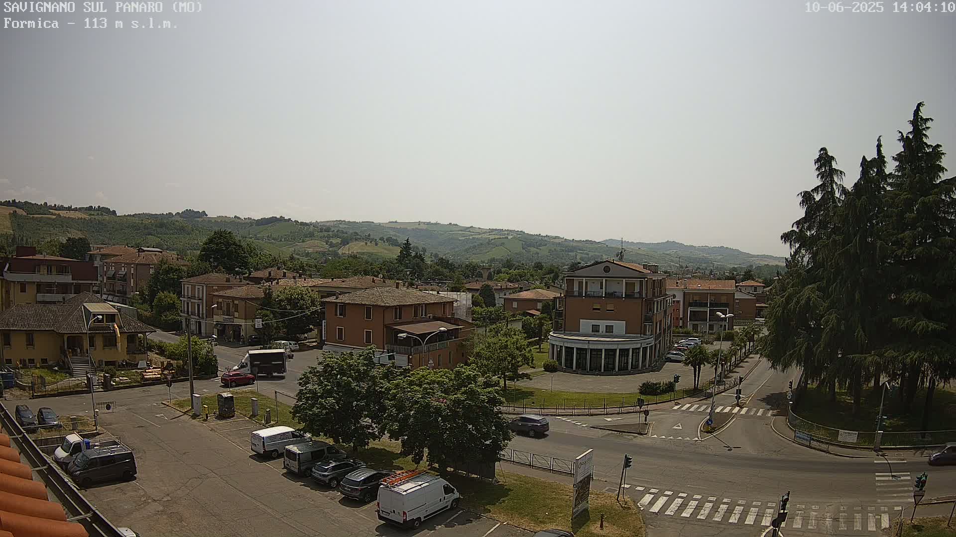 A sunny day reveals a high-angle view of a town square nestled amongst low hills, with several buildings, vehicles, and trees visible.