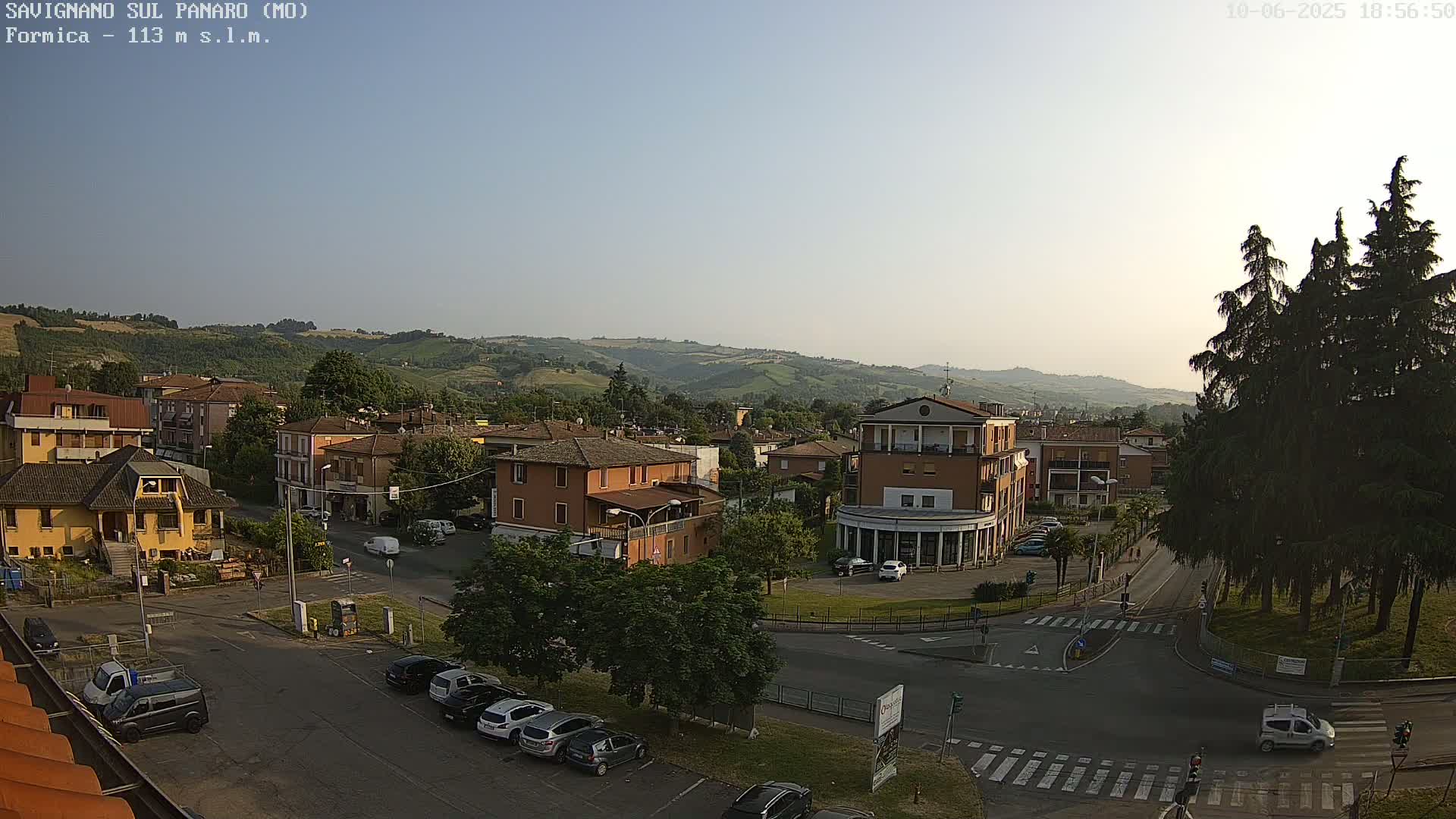 A clear, late afternoon view of a town nestled in a valley, surrounded by rolling green hills, with cars parked along streets and trees lining the edges.