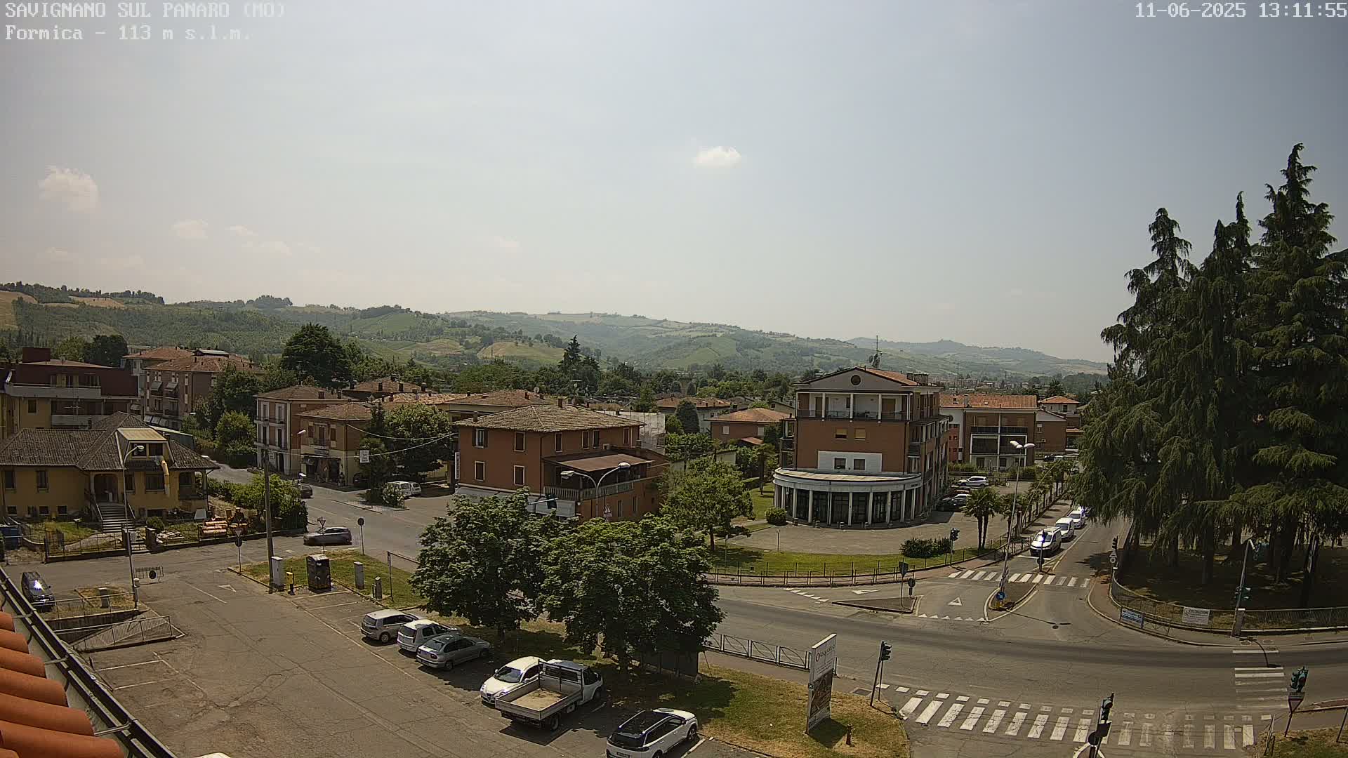 A sunny, high-angle view of a town square with buildings, parked cars, and a few cars driving on roads, surrounded by green hills in the distance.