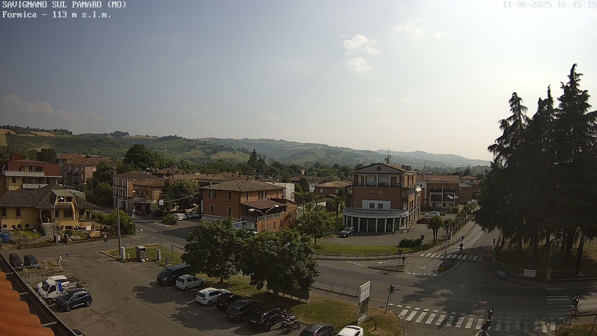A partly sunny view of a town nestled in a hilly landscape, with cars parked in a lot and several buildings visible.