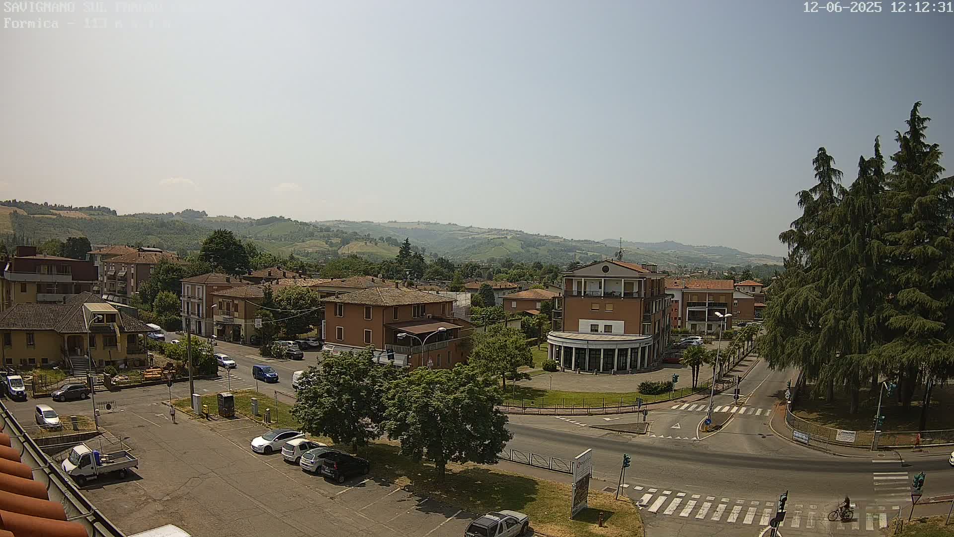 A sunny day reveals a town square in a small Italian town, surrounded by low-rise buildings and lush green hills in the background.