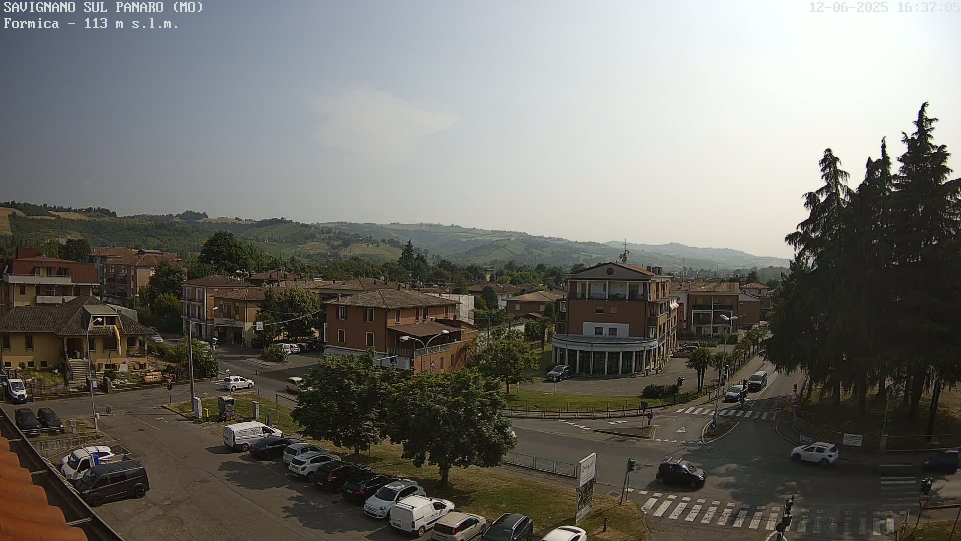 A high-angle, sunny view of a town square with buildings, cars, and trees, set against a backdrop of rolling hills.