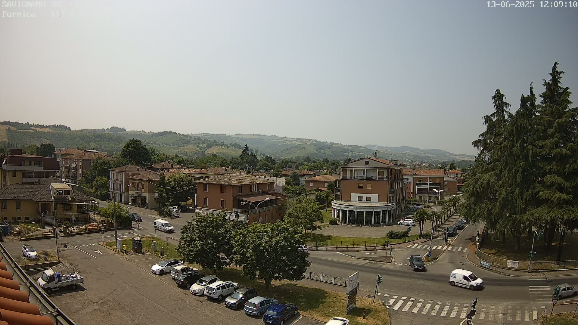 A high-angle, sunny view of a town square with several buildings, cars, and green spaces, set against a backdrop of rolling hills.