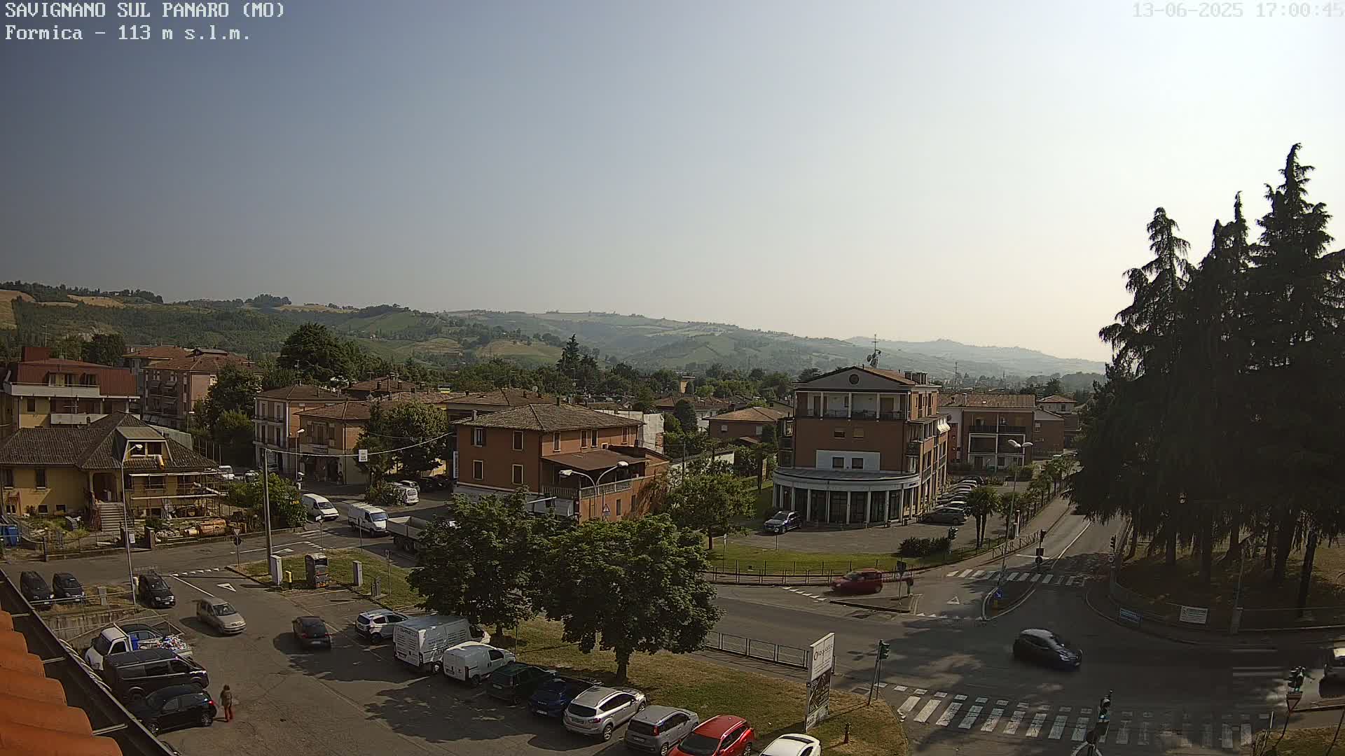 A sunny day reveals a panoramic view of a town nestled in a valley, with cars parked along streets and a few driving through intersections.