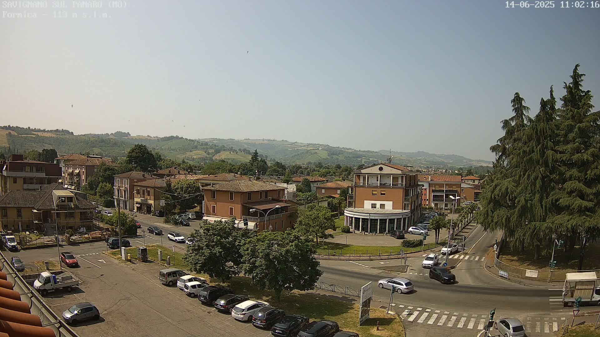 A sunny day reveals a town square with parked cars and buildings, set against a backdrop of rolling hills.