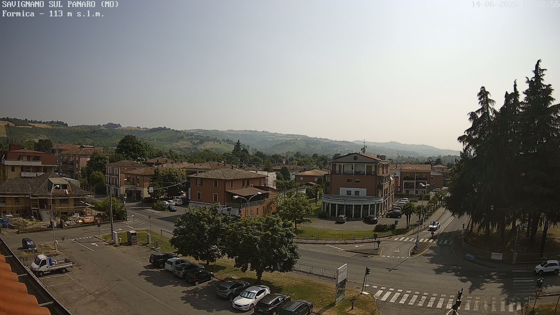 A sunny, hazy day reveals a town nestled in a valley, with rolling hills in the background and cars parked in a paved lot in the foreground.