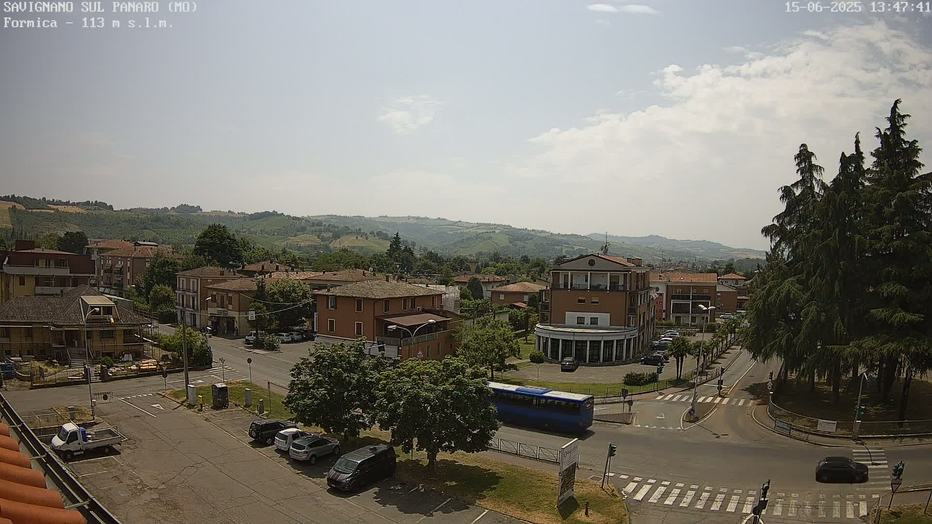 A partly sunny view of a town square, with buildings, cars, a bus, and trees, situated in a valley with rolling hills in the background.
