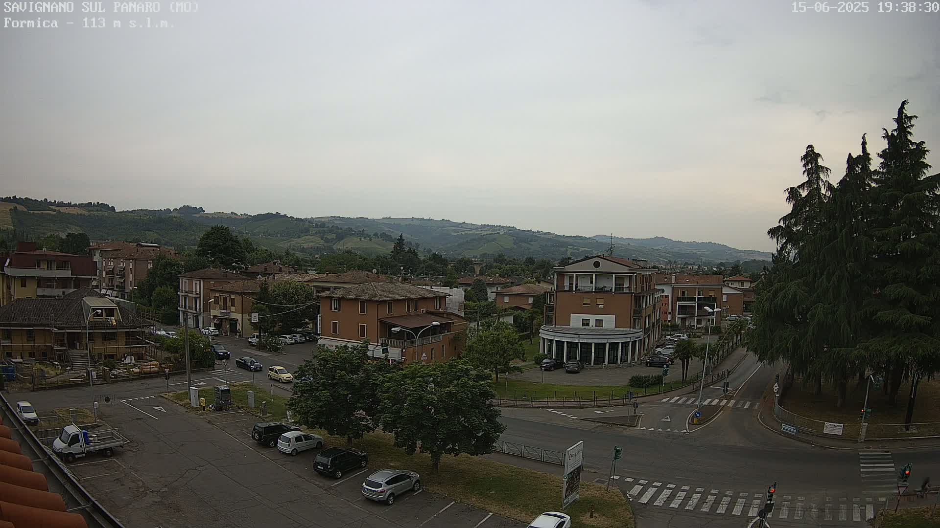 A high-angle, long shot of a town square with cars parked and driving, under a cloudy sky, backed by rolling green hills.
