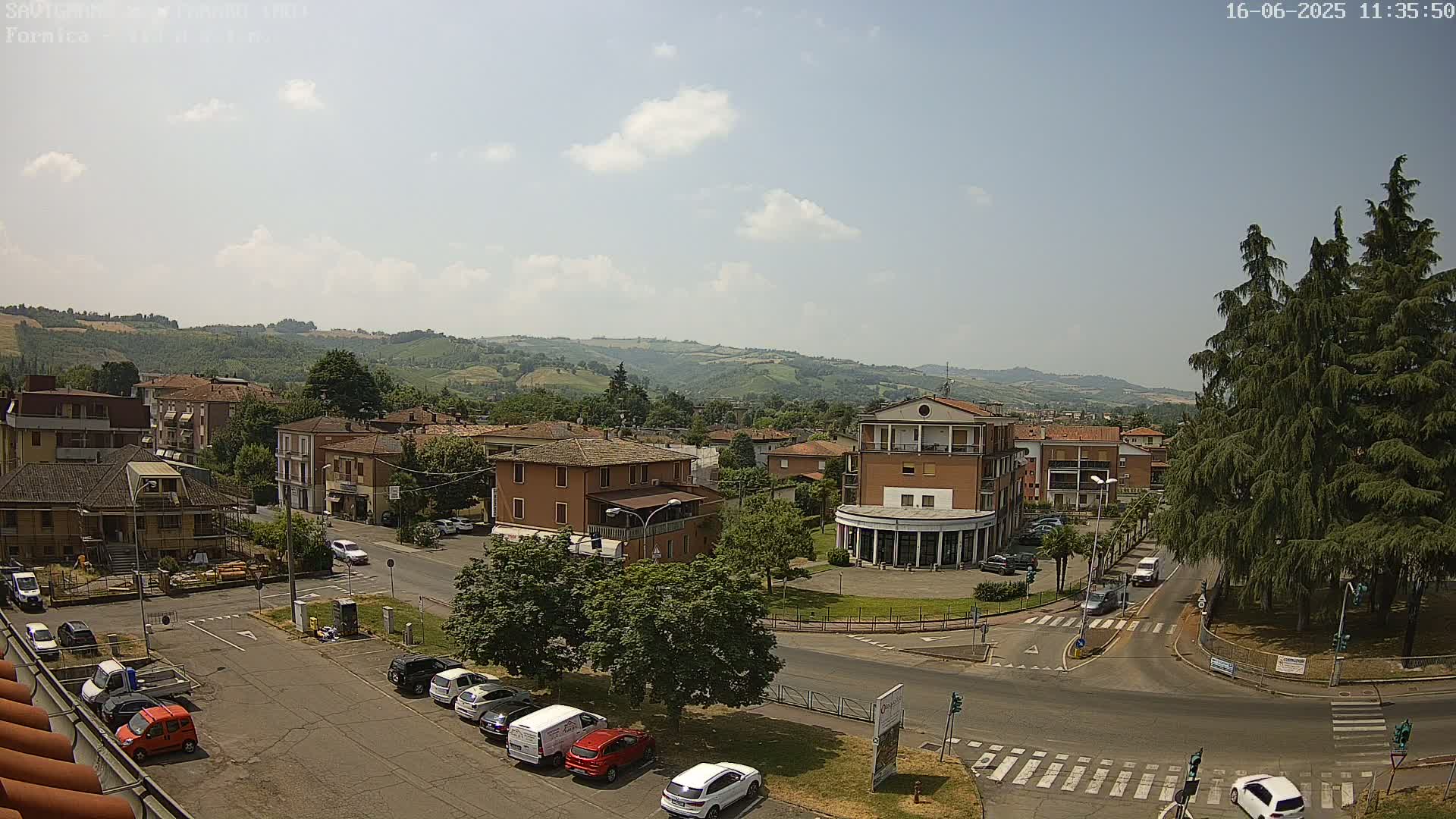 A sunny day reveals a town square with parked cars, several buildings, and rolling hills in the background.