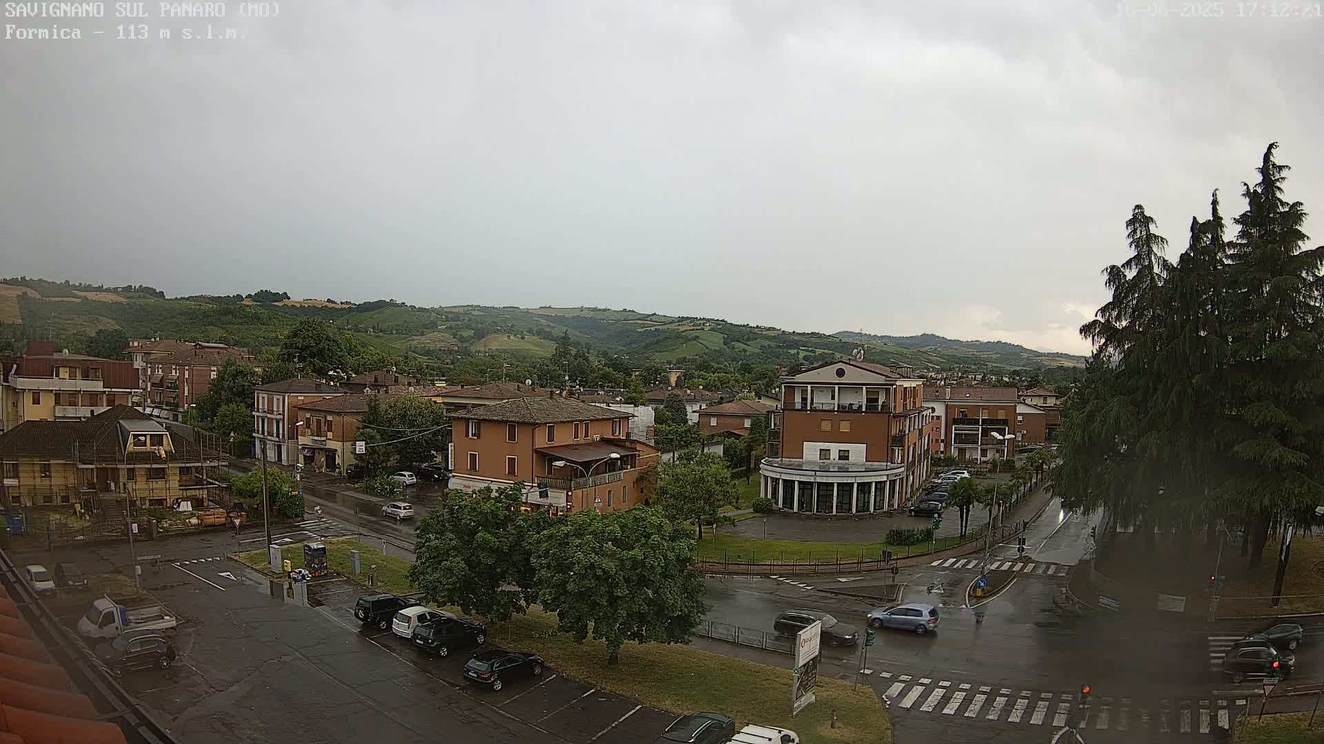 A high-angle, long shot of a town square in the rain, with several buildings, cars, and green spaces, situated against a backdrop of rolling hills under an overcast sky.