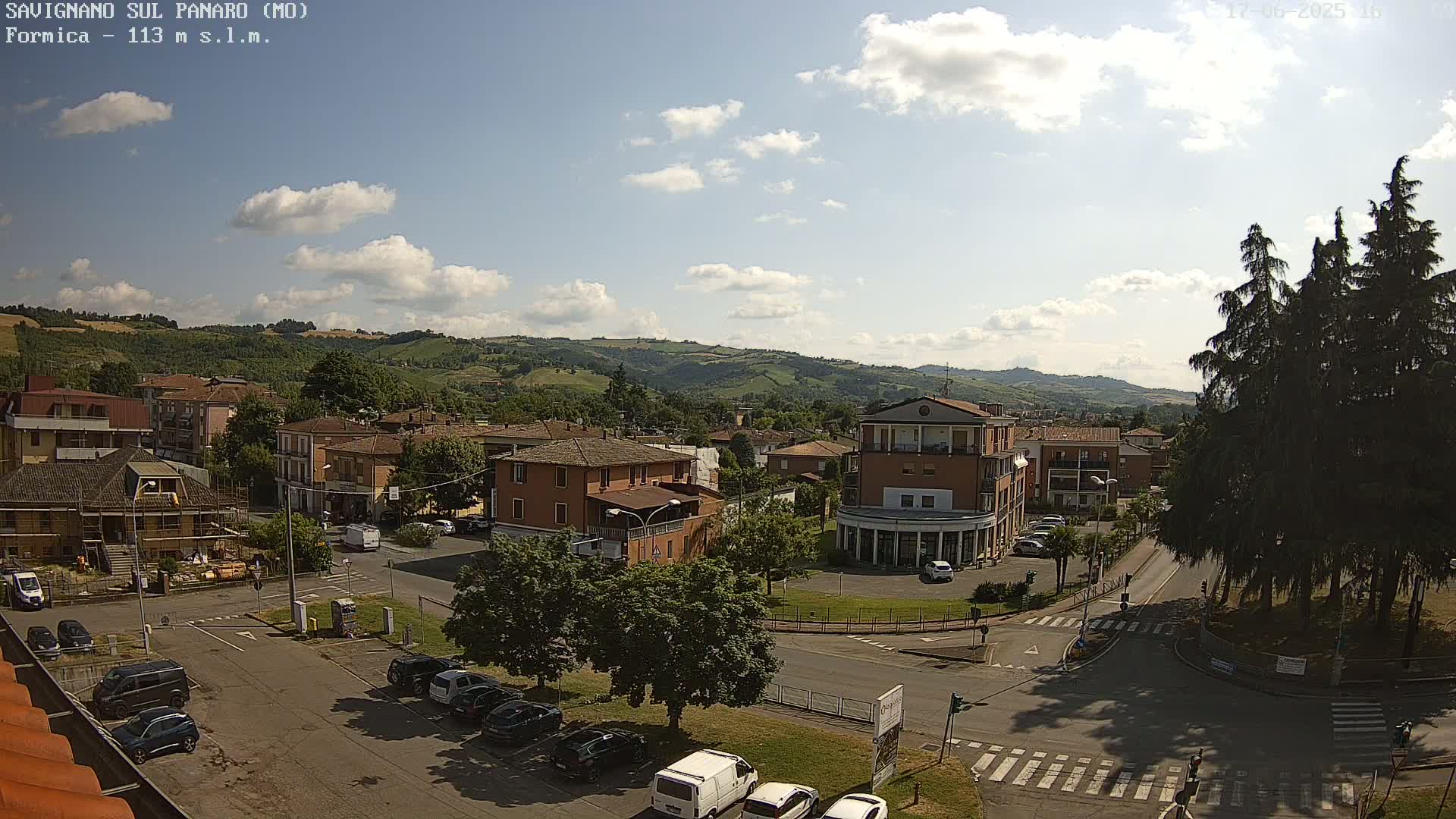 A partly sunny view of a town square and surrounding buildings nestled in a hilly landscape, with cars parked along the streets and trees lining the square.