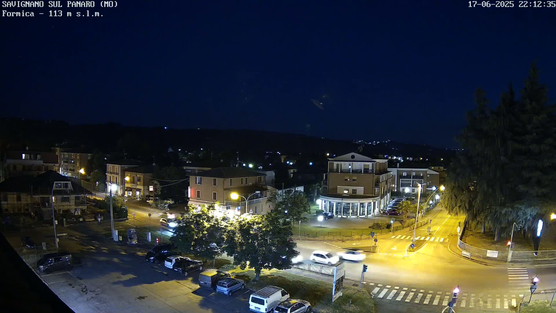 A nighttime aerial view of a town square with cars parked and driving on the roads under a clear, dark sky.