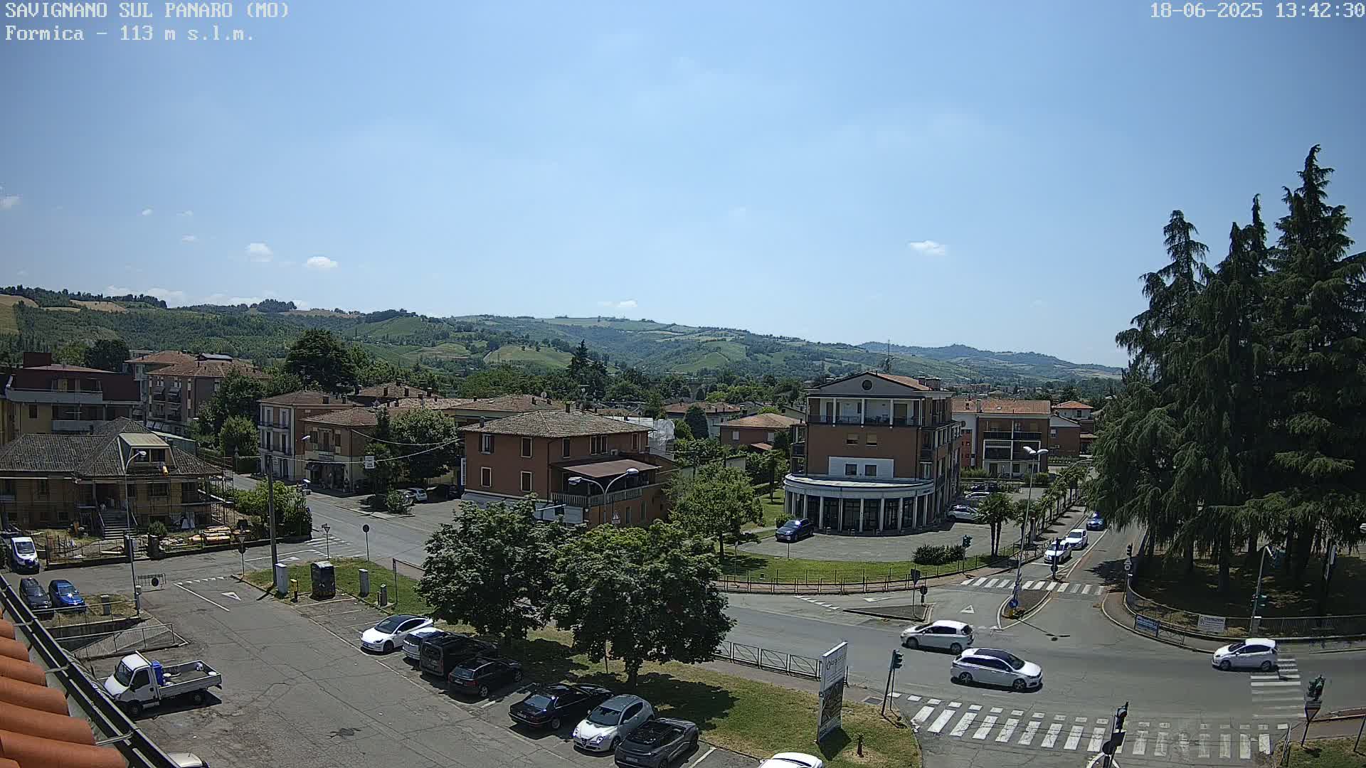 A sunny day reveals a town nestled in a valley, with cars driving on roads that wind through buildings and parking lots, all overlooked by rolling green hills.