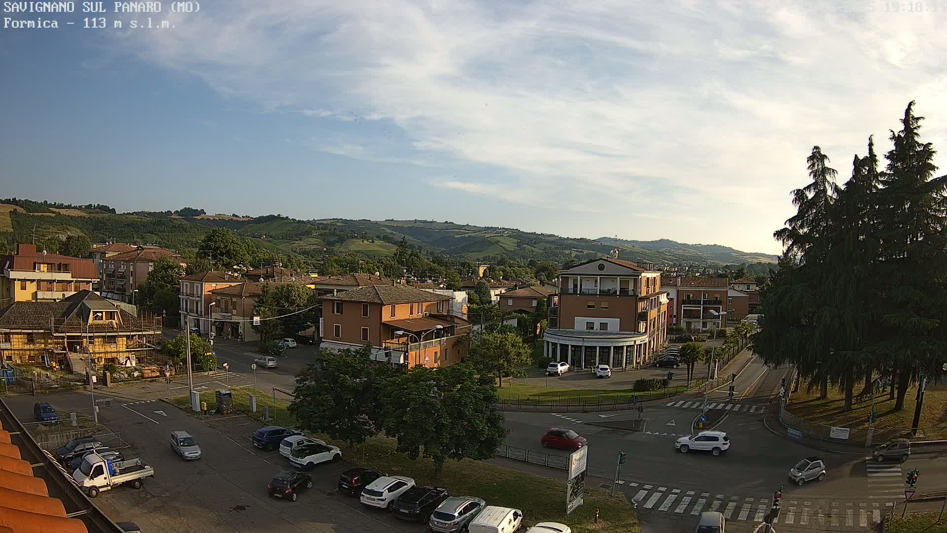 An aerial view of a town nestled in a valley under a partly cloudy sky shows several buildings, parked cars, and a few cars driving on the roads.