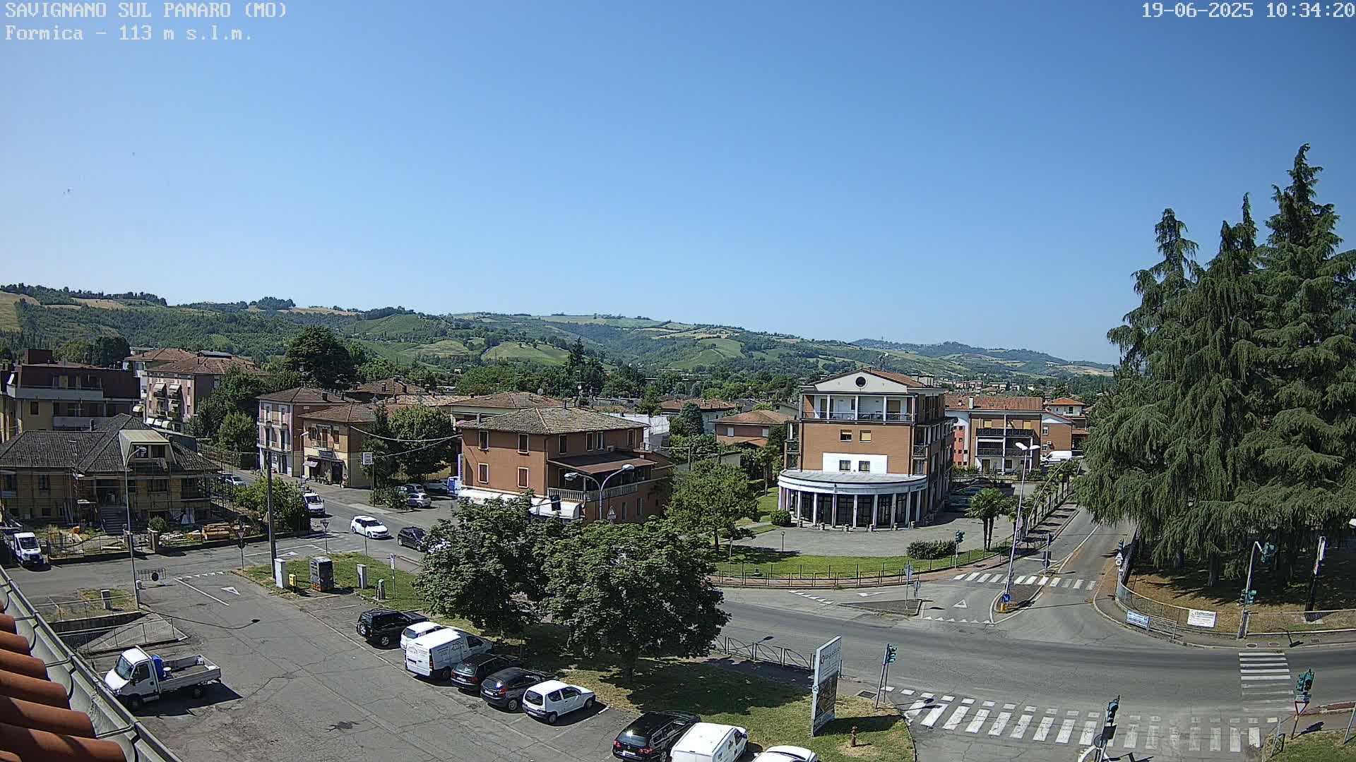 A sunny, high-angle view of a town square with several buildings, cars, and lush green trees, all set against a backdrop of rolling hills.