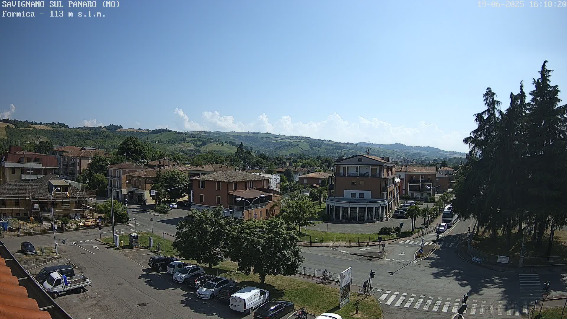 A sunny day reveals a small town nestled in a valley, surrounded by rolling hills and dotted with parked cars and trees.