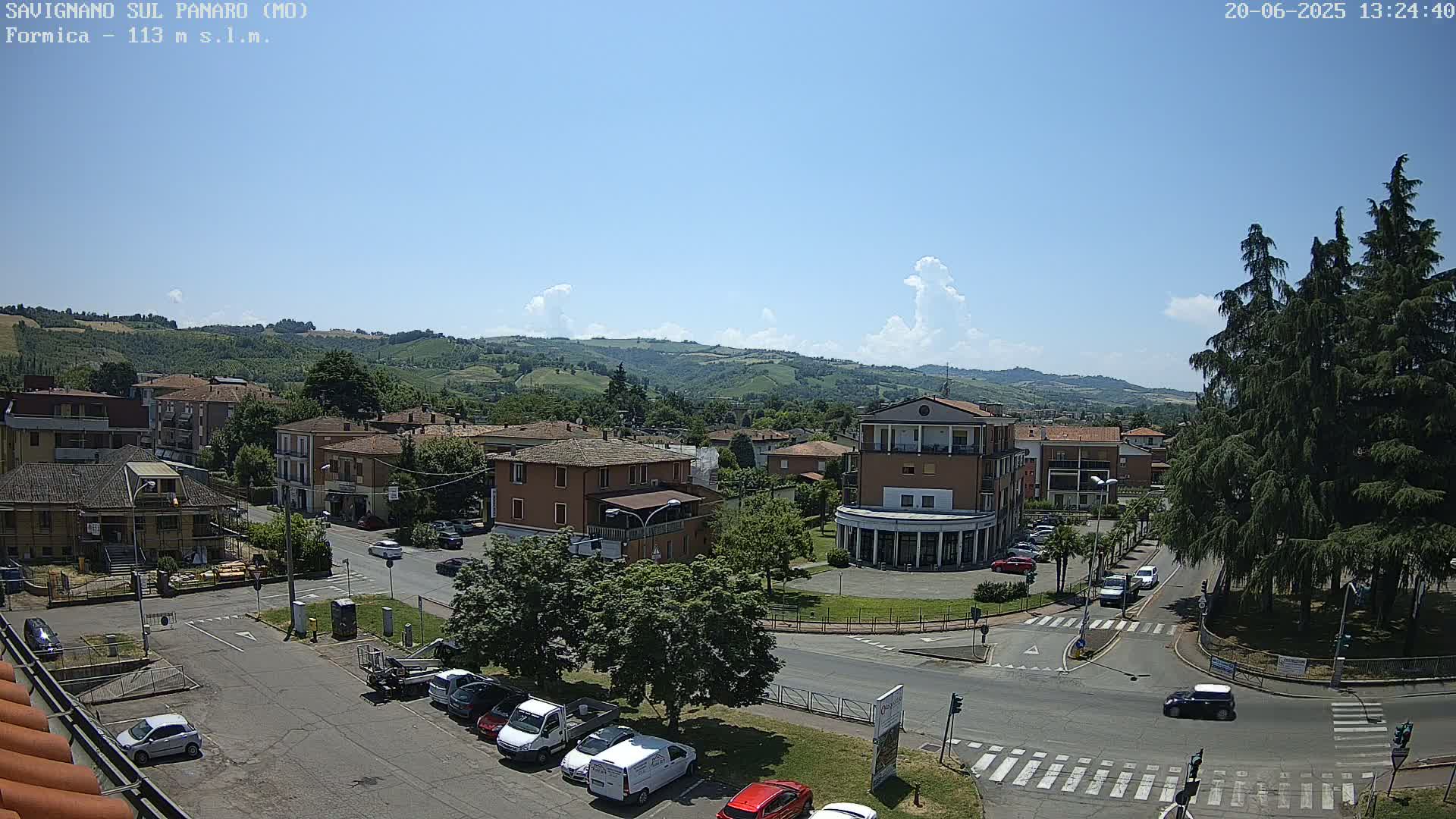 A sunny day reveals a view of a town nestled in rolling green hills, with cars parked in a lot and roads intersecting near a circular area with tall trees.