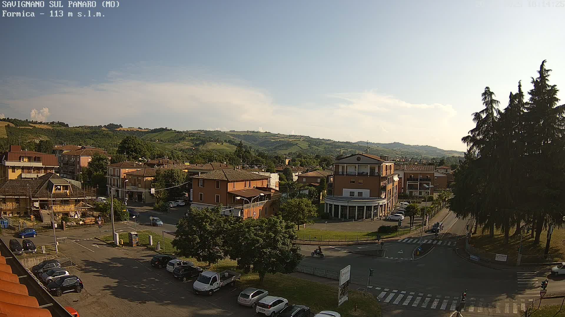 A partly cloudy day reveals a small town nestled in a valley, surrounded by rolling hills and a few evergreen trees.